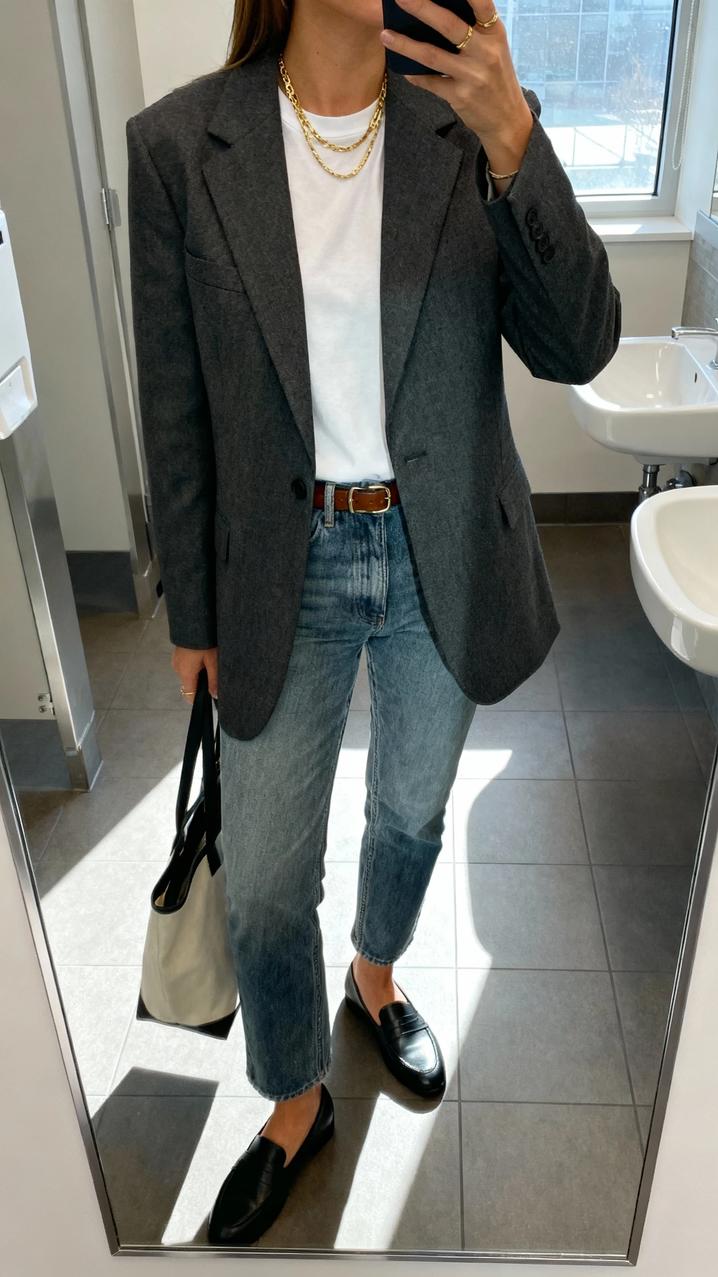 iPhone mirror selfie of a woman in a tailored charcoal blazer over a crisp white tee, medium-wash straight jeans, black loafers, thin leather belt, layered gold necklaces, and a tote bag, face not visible, office restroom mirror, soft overhead lighting with window spill, natural iPhone aesthetic.