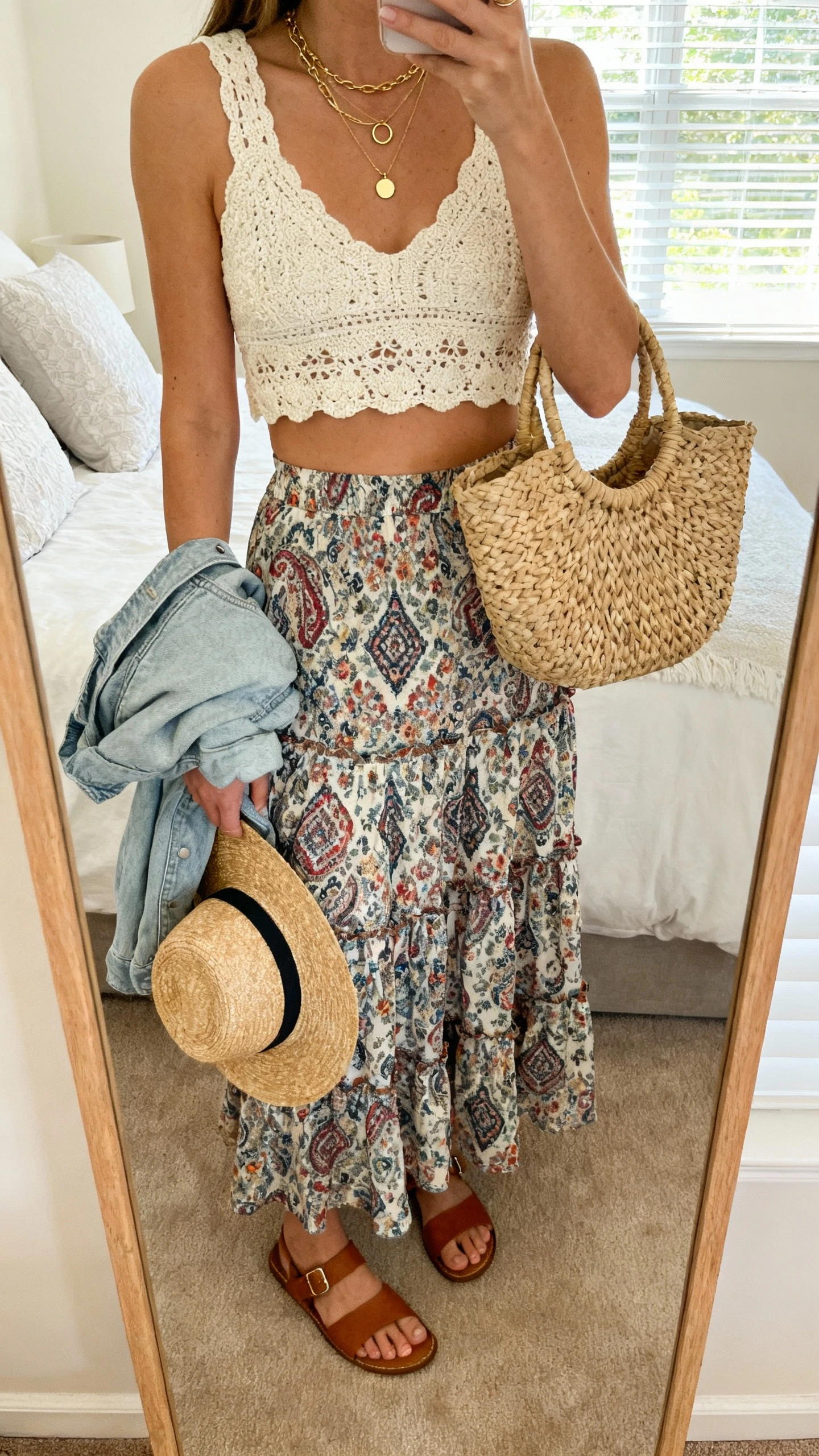 iPhone mirror selfie of a woman in a tiered printed maxi skirt (floral/paisley), cream crochet crop top, flat leather sandals, layered gold necklaces with small hoops, woven straw tote, straw sun hat, optional lightweight denim jacket draped on arm, face not visible, bedroom mirror, morning window light, natural iPhone photo quality.