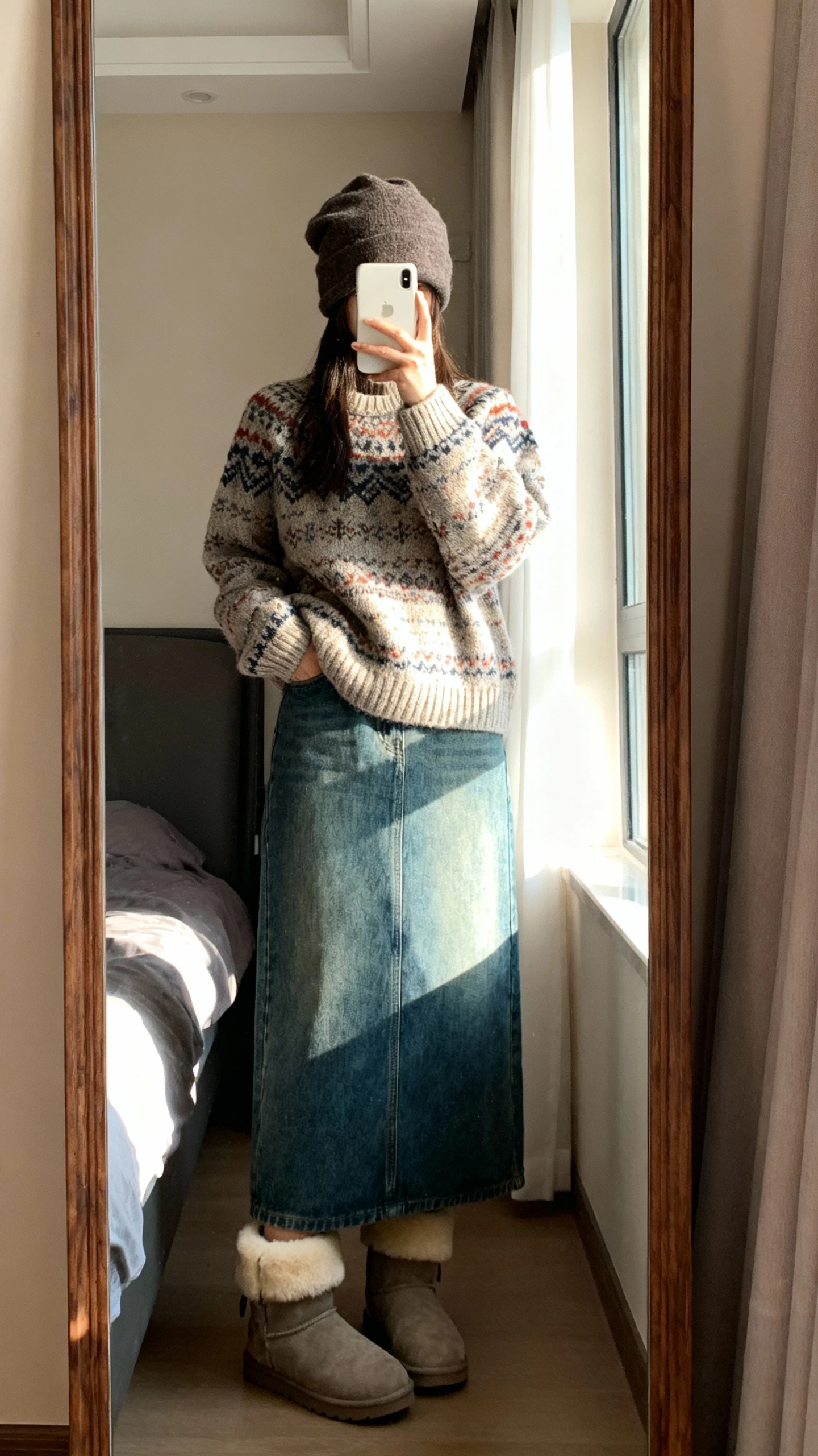 iPhone mirror selfie of a woman in an artisan knit sweater, denim maxi skirt, and shearling boots with a slouchy beanie, face not visible, bedroom full-length mirror, daylight from window, natural iPhone photo quality.