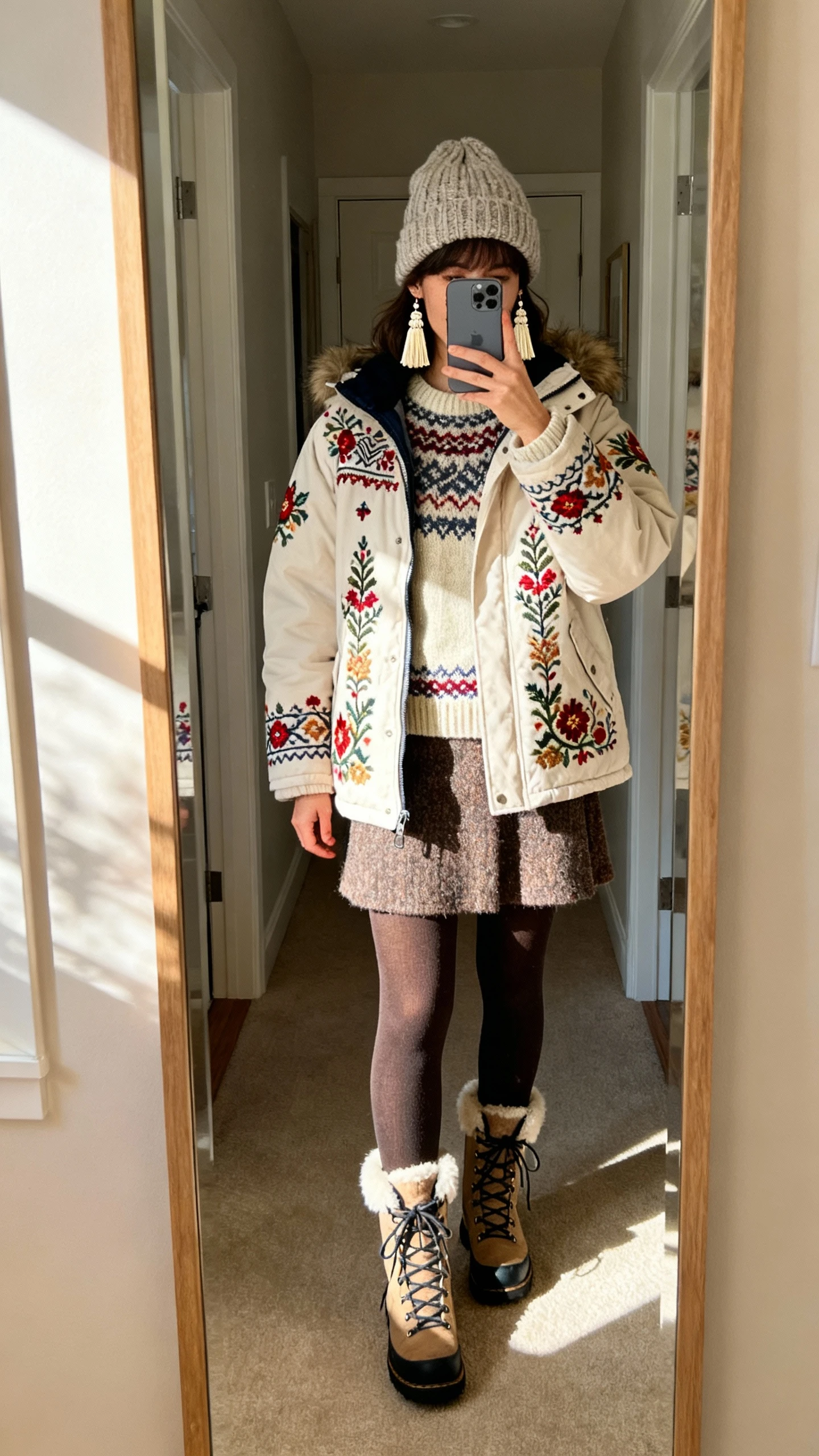 iPhone mirror selfie of a woman in an embroidered parka with folklore motifs, fair isle sweater, wool skirt with tights, lace-up winter boots, knit beanie, and tassel earrings, face not visible, entryway mirror with afternoon window light, natural iPhone photo quality.