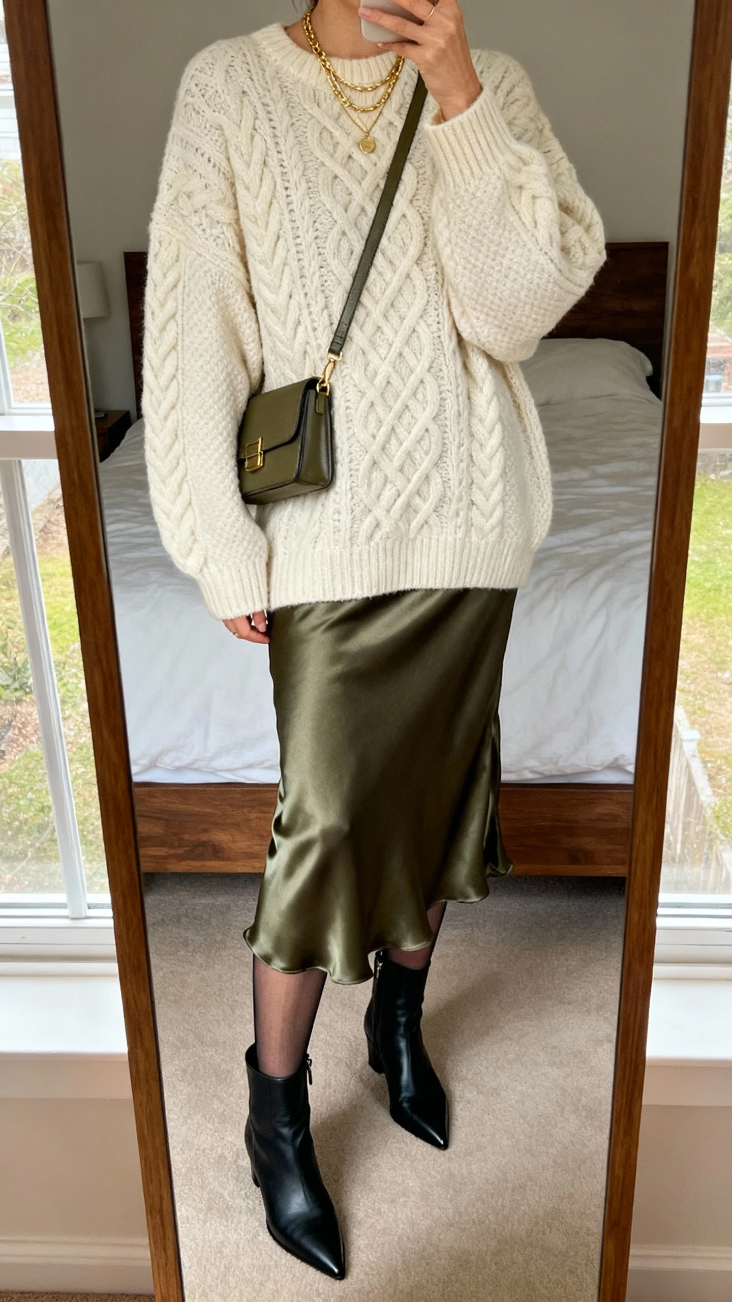 iPhone mirror selfie of a woman in an oversized cream cable-knit sweater, olive satin midi slip skirt, black pointed-toe ankle boots, sheer tights, delicate gold layered necklaces, and a structured crossbody bag; face not visible, cropped at neck, bedroom mirror with morning window light, natural iPhone photo quality.