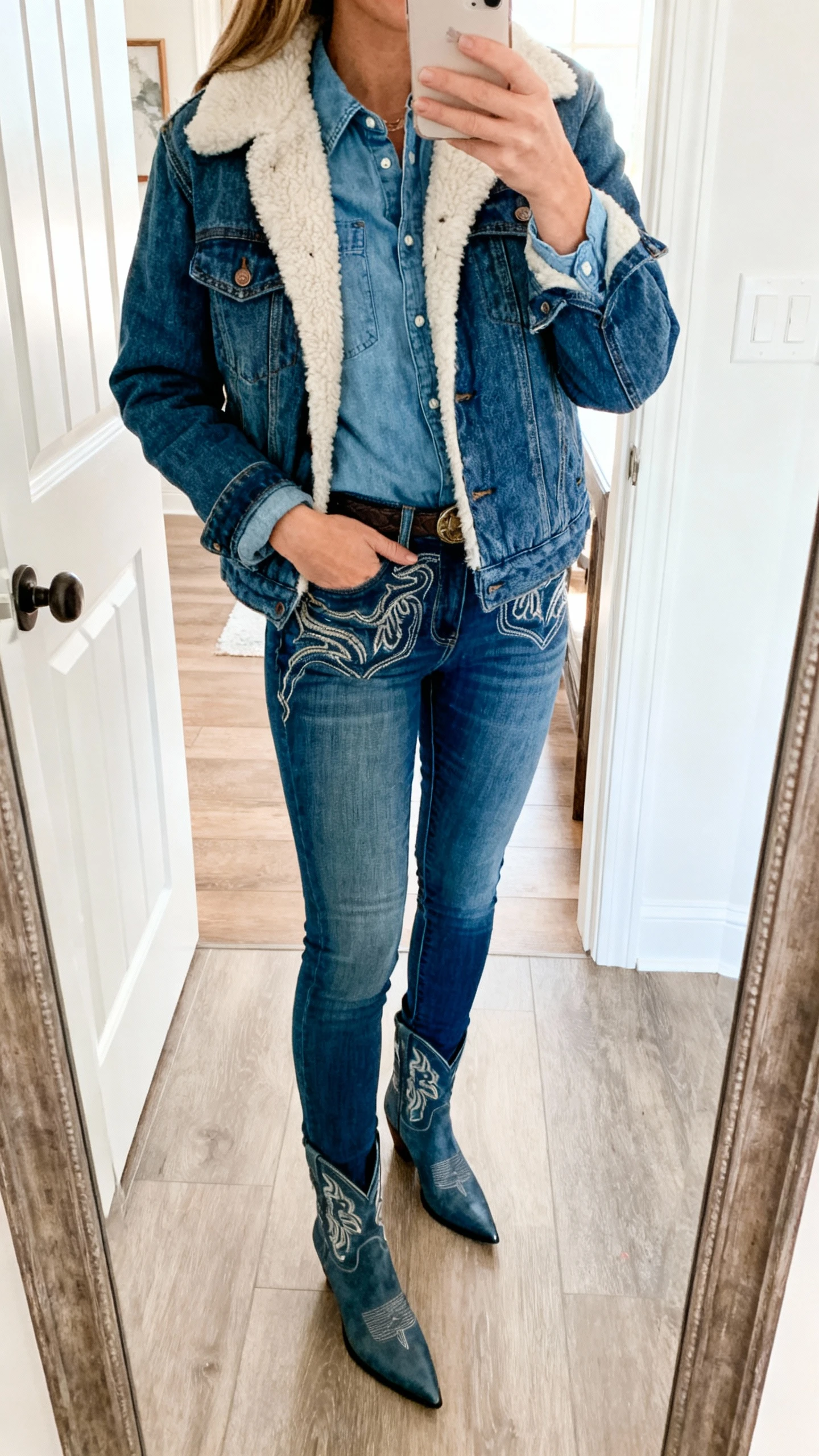 iPhone mirror selfie of a woman in double denim: sherpa-lined trucker jacket over a chambray shirt with western-style skinny jeans and pointed-toe boots; face not visible, entryway mirror, bright natural light, casual iPhone look.