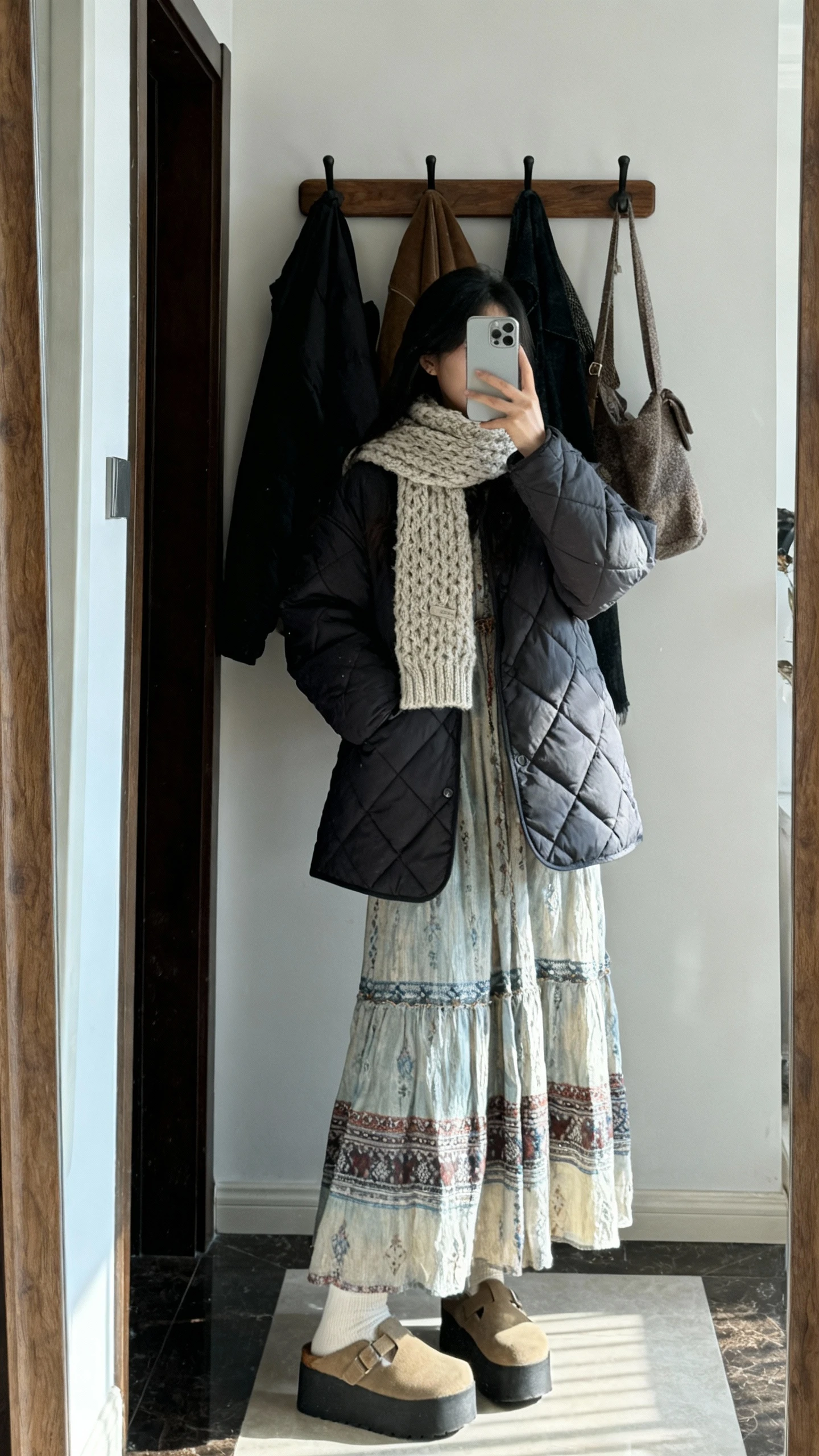 iPhone mirror selfie of a woman wearing a boho maxi dress with a quilted coat and platform clogs, knit scarf, face not visible, entryway mirror with coat rack, cool afternoon light, natural iPhone photo quality.