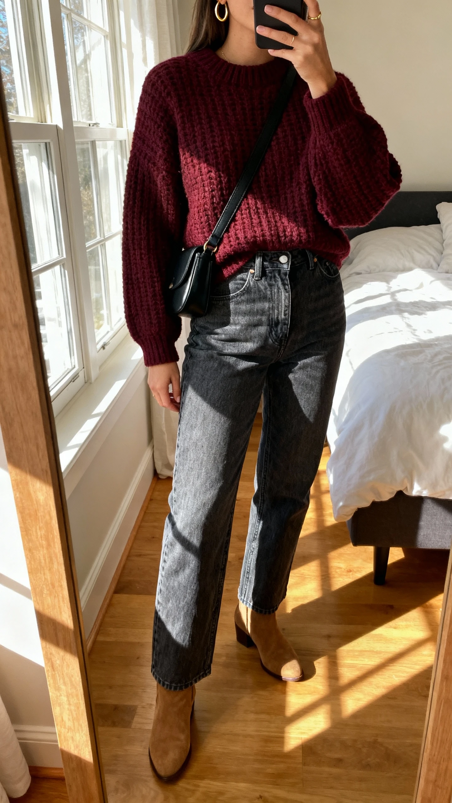 iPhone mirror selfie of a woman wearing a burgundy chunky sweater, high-rise medium-wash straight jeans, tan ankle boots, simple gold hoops, and a black crossbody, face not visible, cropped at neck, bedroom mirror with morning window light, natural iPhone photo quality.