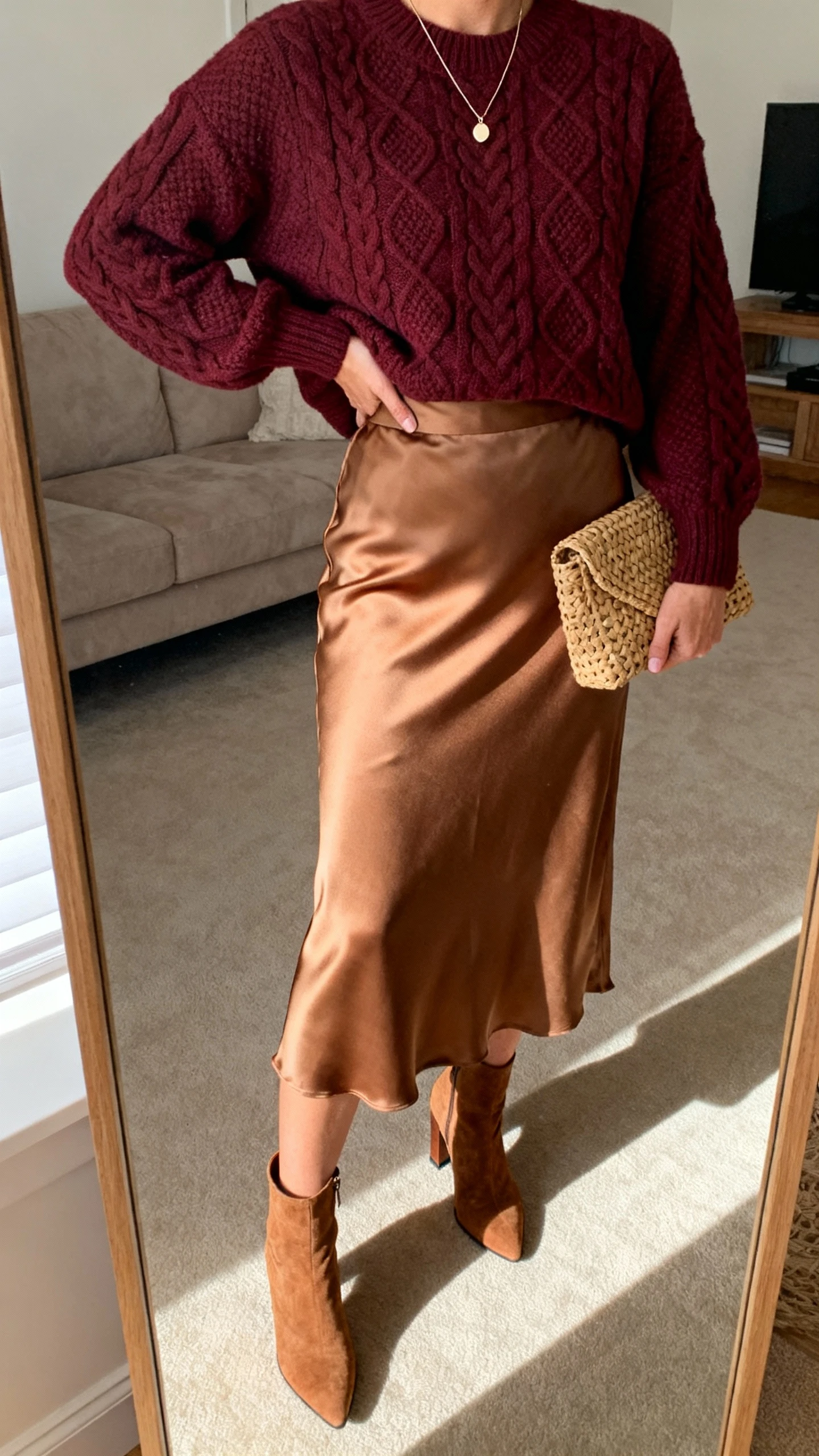 iPhone mirror selfie of a woman wearing a burgundy cable-knit sweater tucked into a tan satin midi skirt, heeled ankle boots, dainty pendant, and a woven clutch, face not visible, cropped at neck, living room mirror, warm indoor lighting, natural iPhone aesthetic.