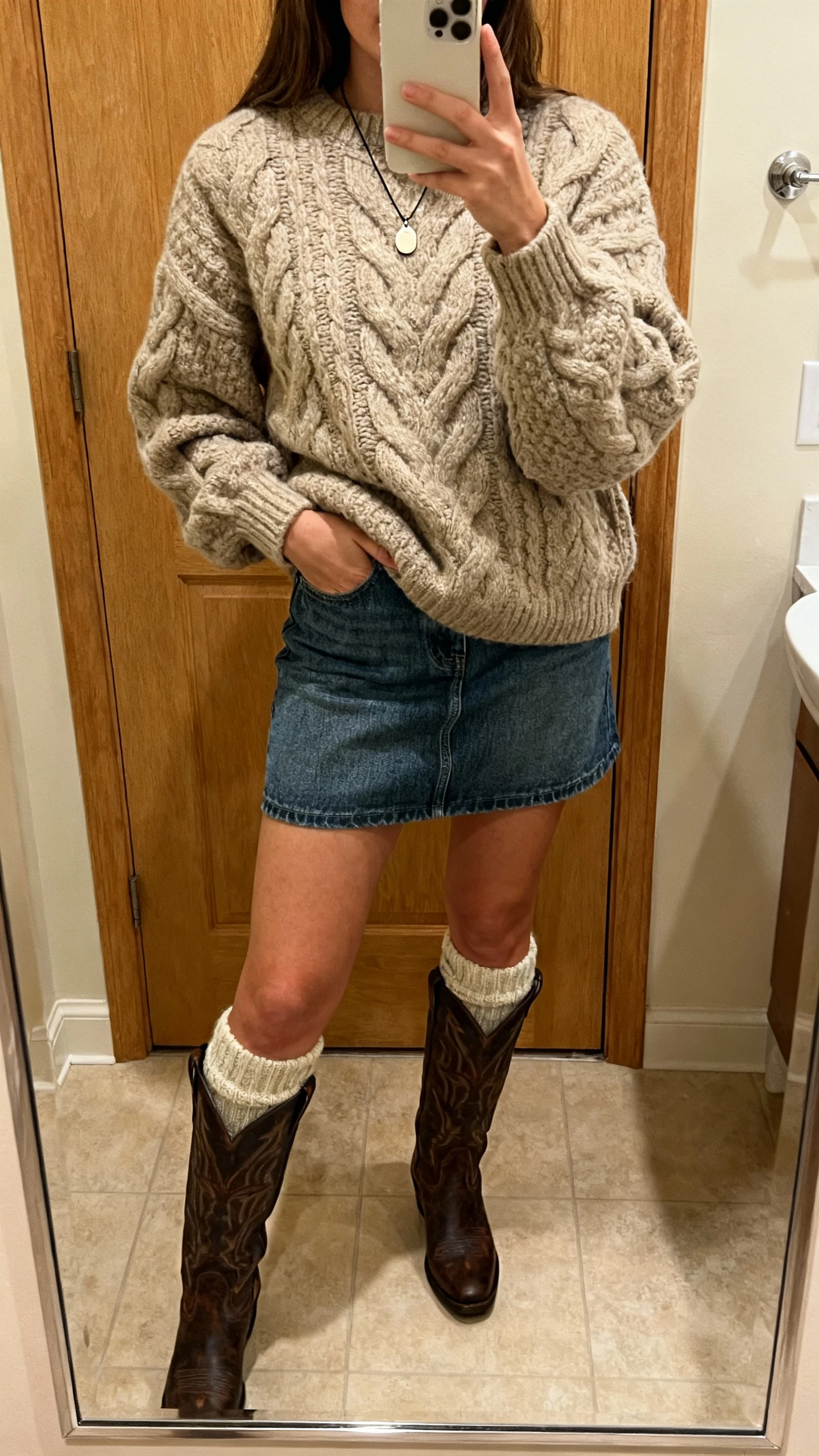 iPhone mirror selfie of a woman wearing a chunky cable-knit sweater tucked into a denim skirt with tall western boots, wool socks peeking, simple pendant necklace; face not visible, bathroom mirror, warm indoor lighting, natural iPhone photo quality.