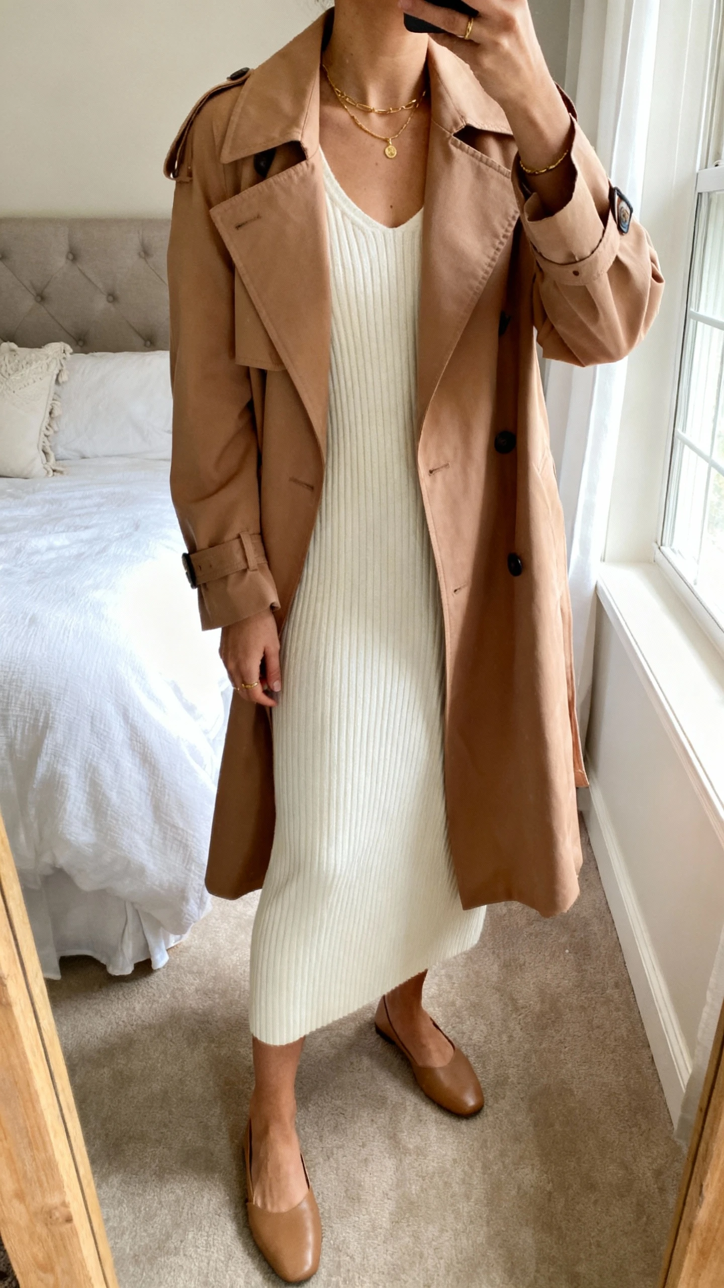 iPhone mirror selfie of a woman wearing a cream knit column dress with a soft camel trench, minimal quiet gold jewelry, and simple leather flats, face not visible, cropped at neck, bedroom mirror with morning window light, natural iPhone photo quality.