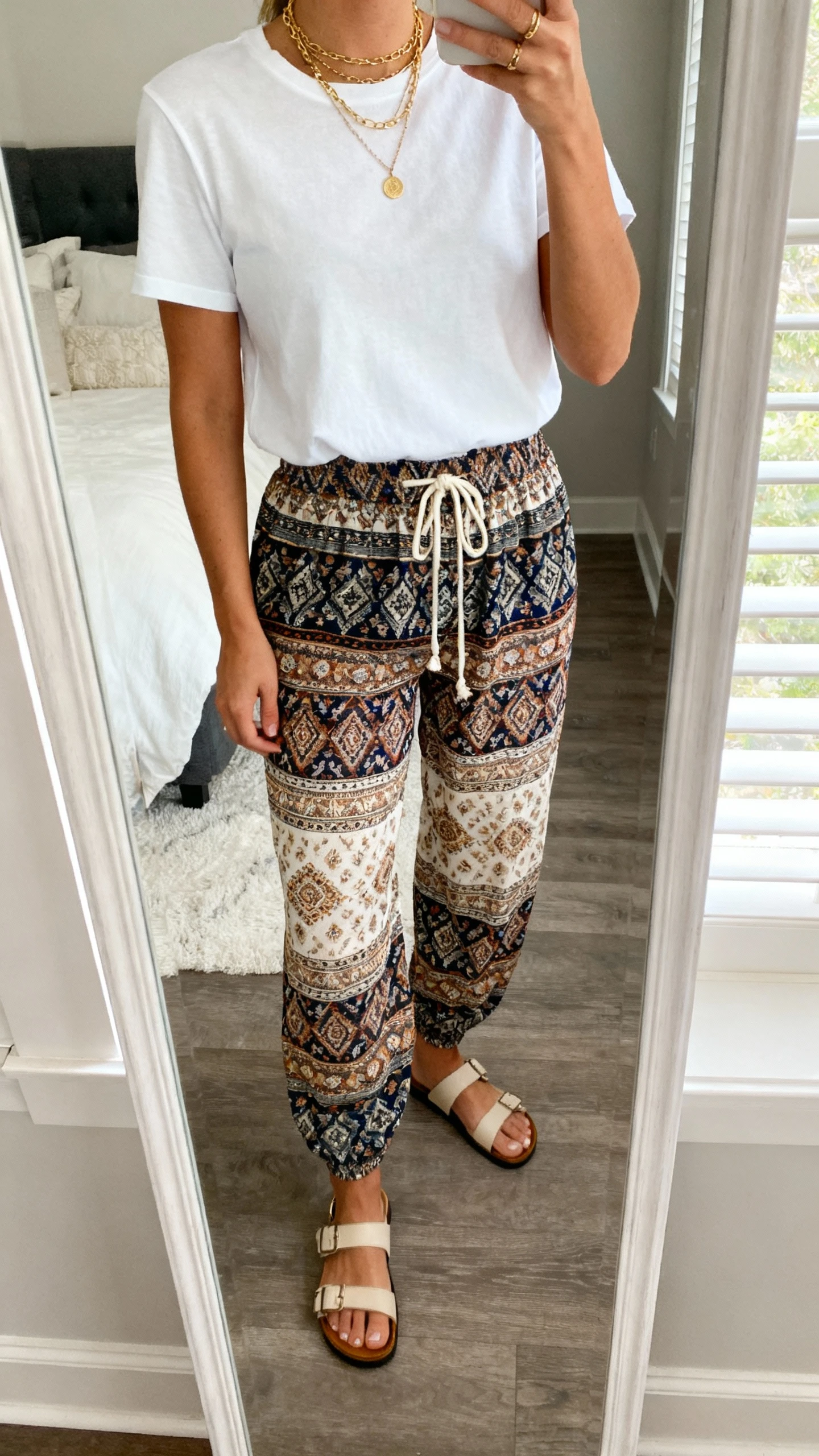 iPhone mirror selfie of a woman wearing a crisp white tee tucked into printed boho pants with a drawstring waist, simple sandals, layered gold necklaces, face not visible, cropped at neck, bedroom mirror with natural window light, natural iPhone photo quality.