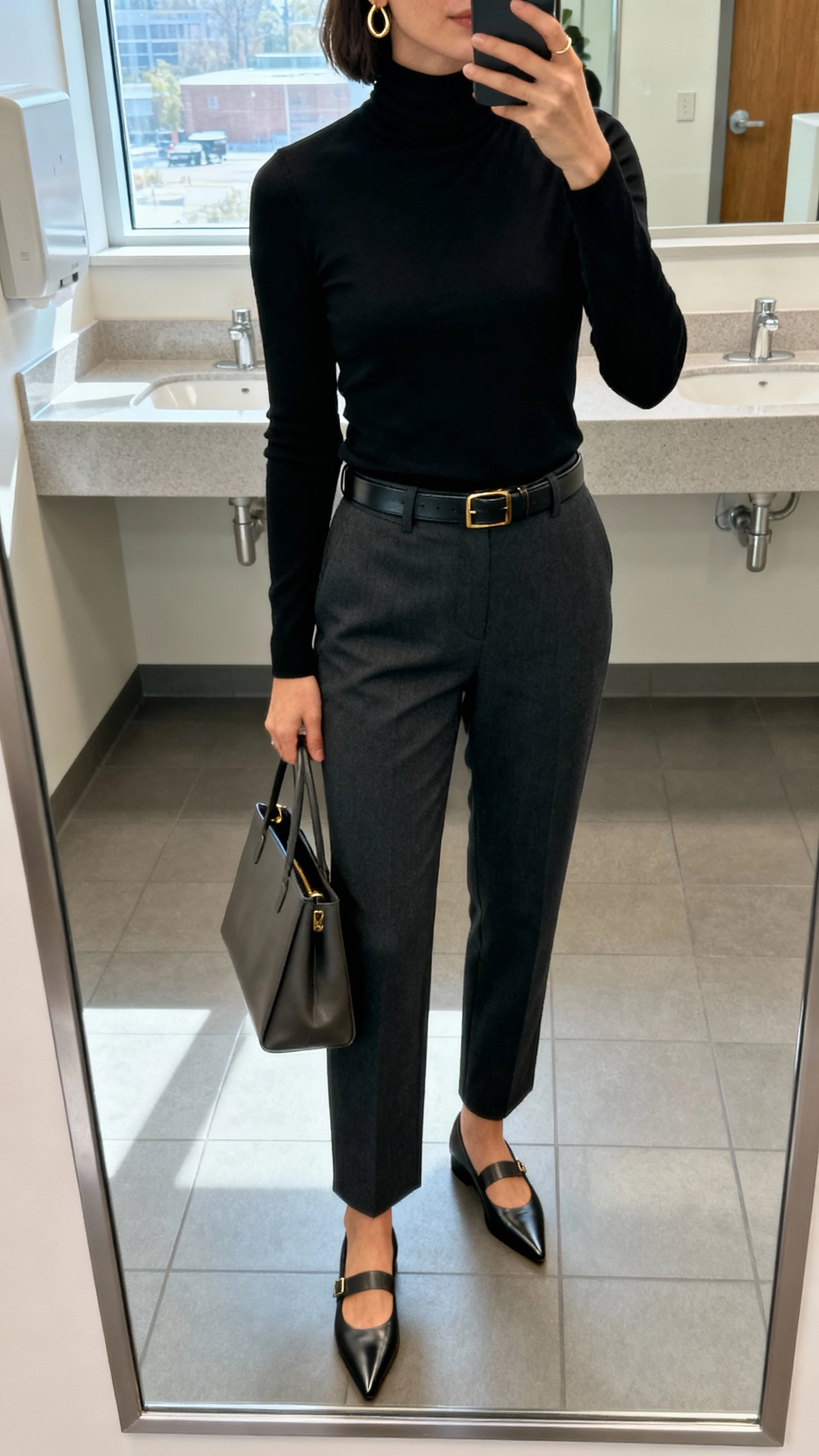 iPhone mirror selfie of a woman wearing a fitted black turtleneck tucked into tailored charcoal trousers, pointed-toe loafers, slim leather belt, minimal gold hoops, structured tote, face not visible, cropped at neck, office restroom mirror, morning window light, natural iPhone photo quality.