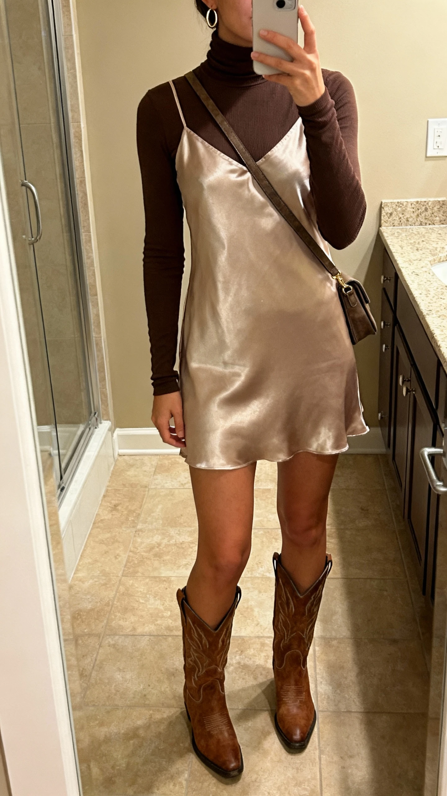 iPhone mirror selfie of a woman wearing a fitted turtleneck under a satin slip dress with Western boots, minimal hoop earrings, crossbody bag, face not visible, bathroom mirror, warm indoor lighting, natural iPhone aesthetic.