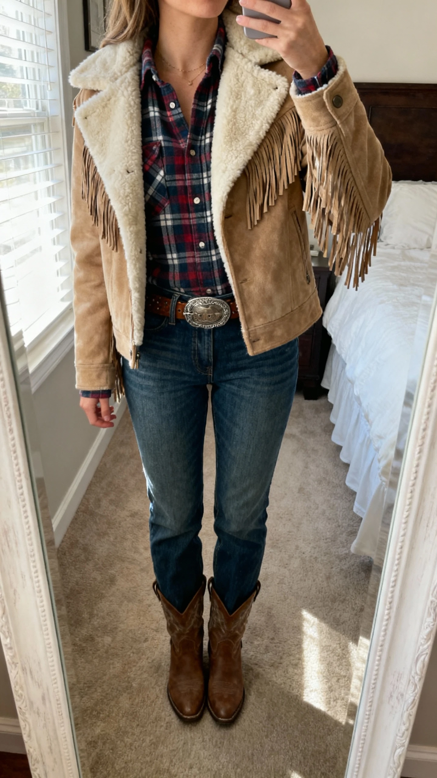 iPhone mirror selfie of a woman wearing a flannel shirt layered under a shearling-lined jacket with fringe details, straight-leg jeans, western belt, and ankle boots; face not visible, cropped at neck, bedroom full-length mirror, soft window light, natural iPhone photo quality.