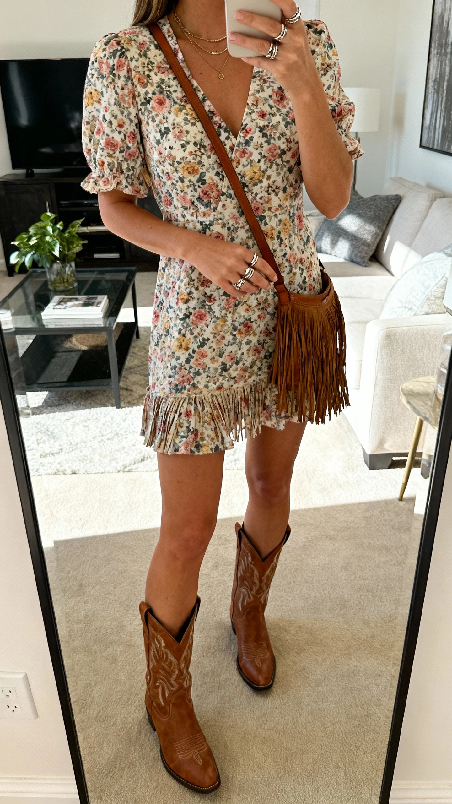 iPhone mirror selfie of a woman wearing a floral mini dress with fringe details, western-style ankle boots, layered rings and a crossbody bag, face not visible, cropped at shoulders, living room mirror, bright daylight, natural iPhone photo.