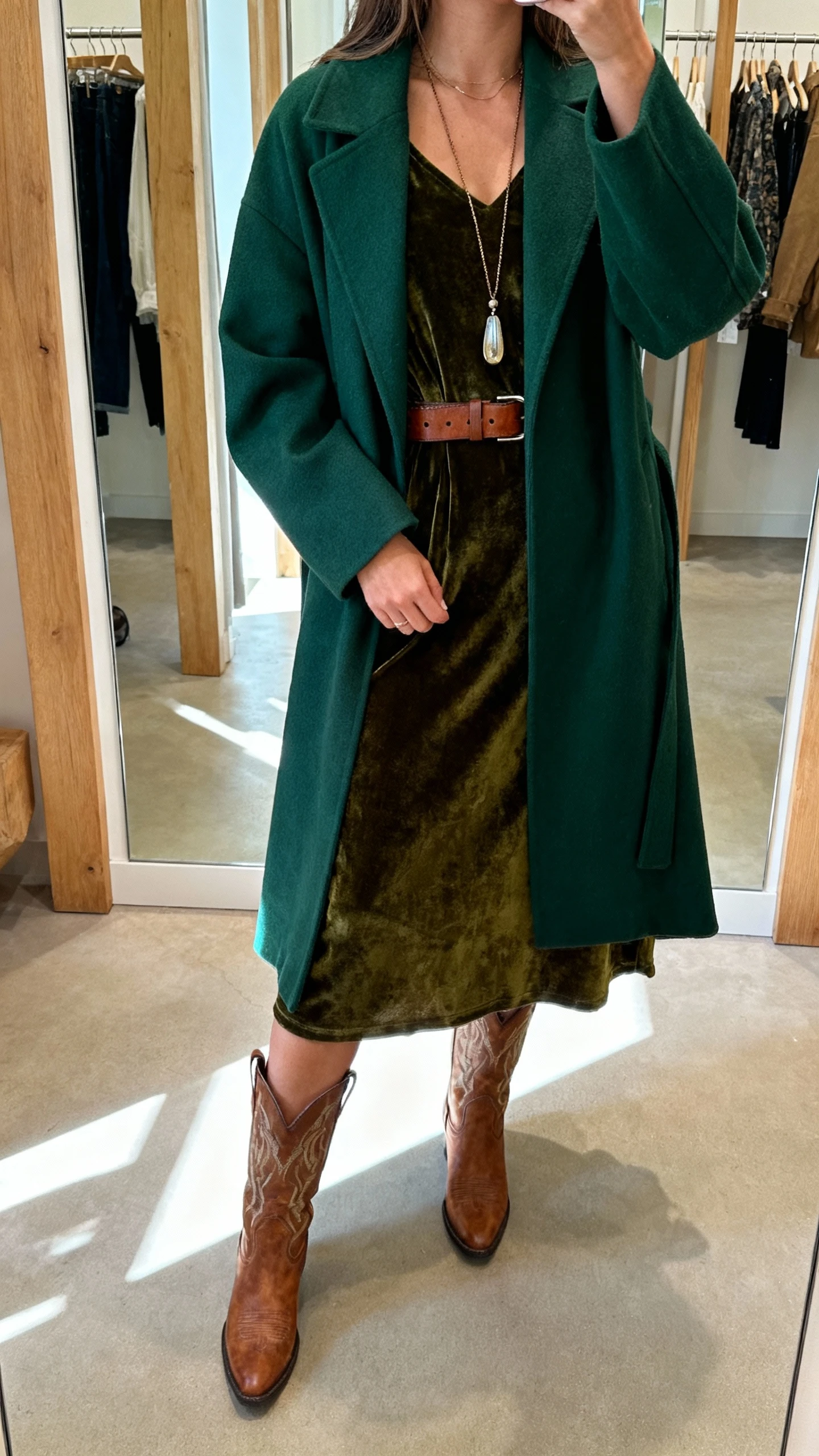 iPhone mirror selfie of a woman wearing a forest-green duster coat over a velvet midi dress, cowboy boots, leather belt, long pendant necklace, face not visible, cropped at neck, boutique dressing-room mirror lighting with soft natural spill, natural iPhone photo quality.