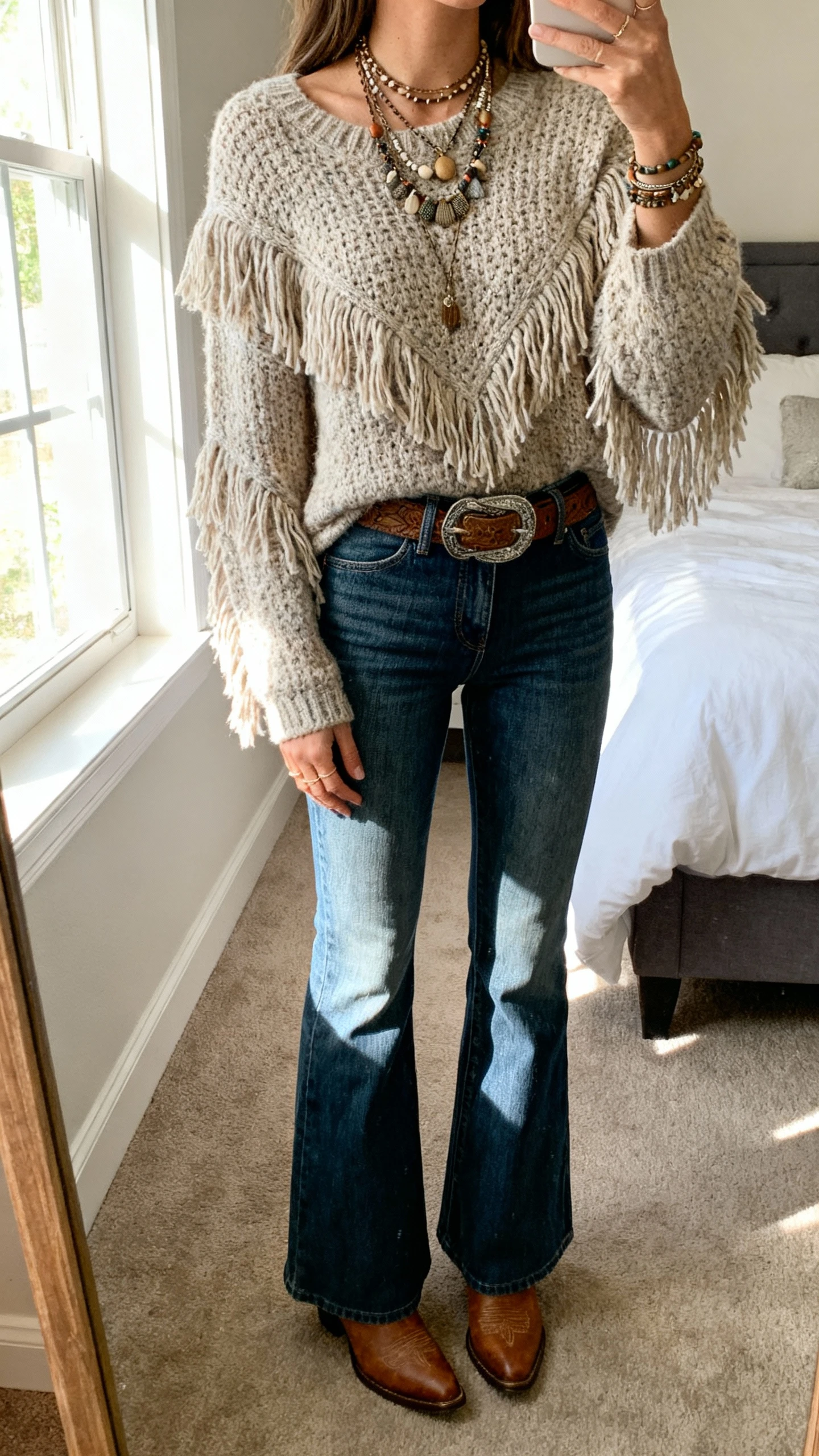iPhone mirror selfie of a woman wearing a fringed knit sweater, high-waisted flare jeans, western belt, and cowboy boots with layered boho jewelry, face not visible, cropped at neck, bedroom mirror with natural window light, natural iPhone photo quality.