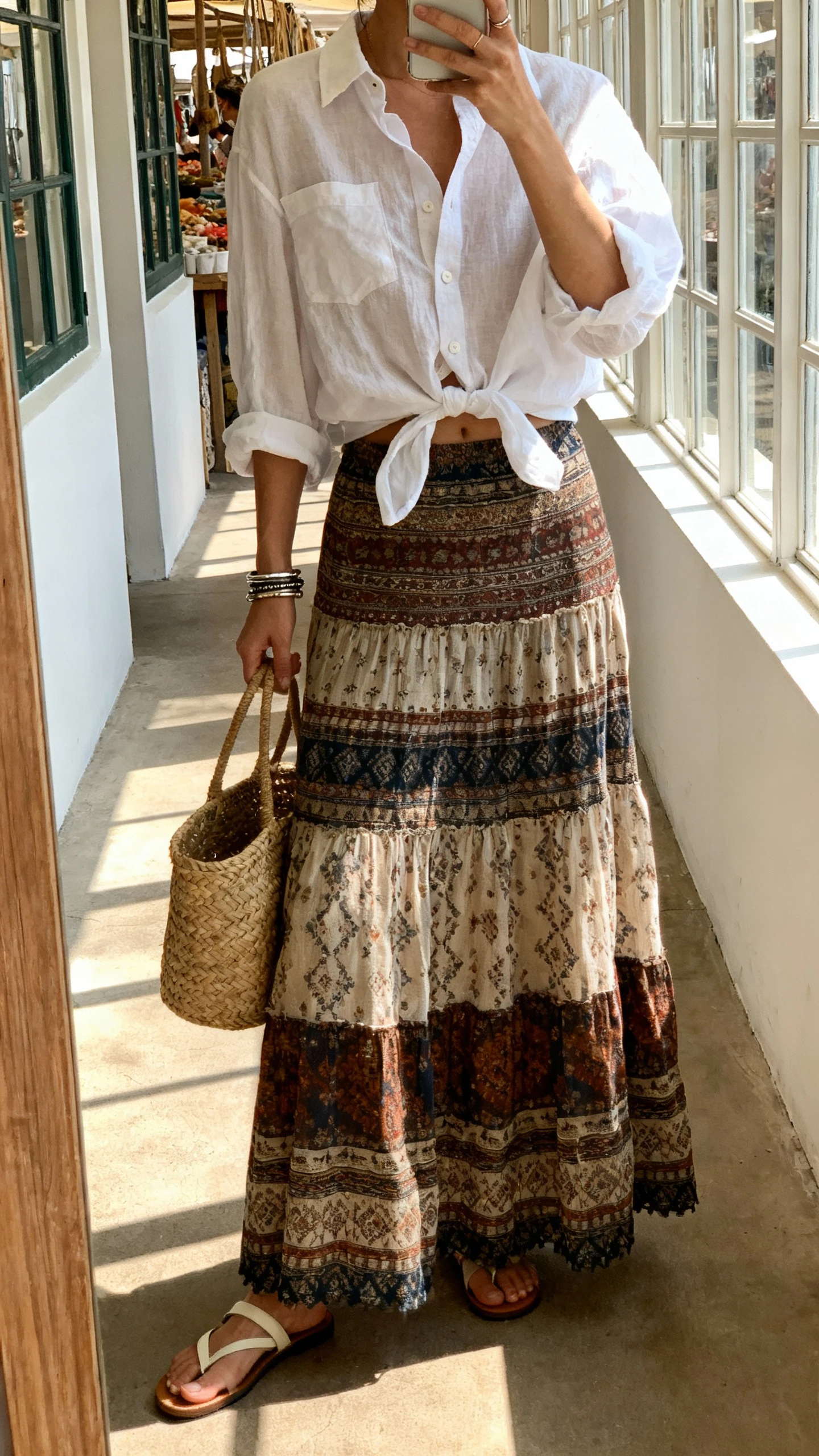 iPhone mirror selfie of a woman wearing a long boho skirt with a breezy white button-down half-tucked, basket bag on arm, simple sandals, layered bracelets, face not visible, cropped at neck, taken in a sunlit hallway like a weekend market morning, natural window light, natural iPhone photo quality