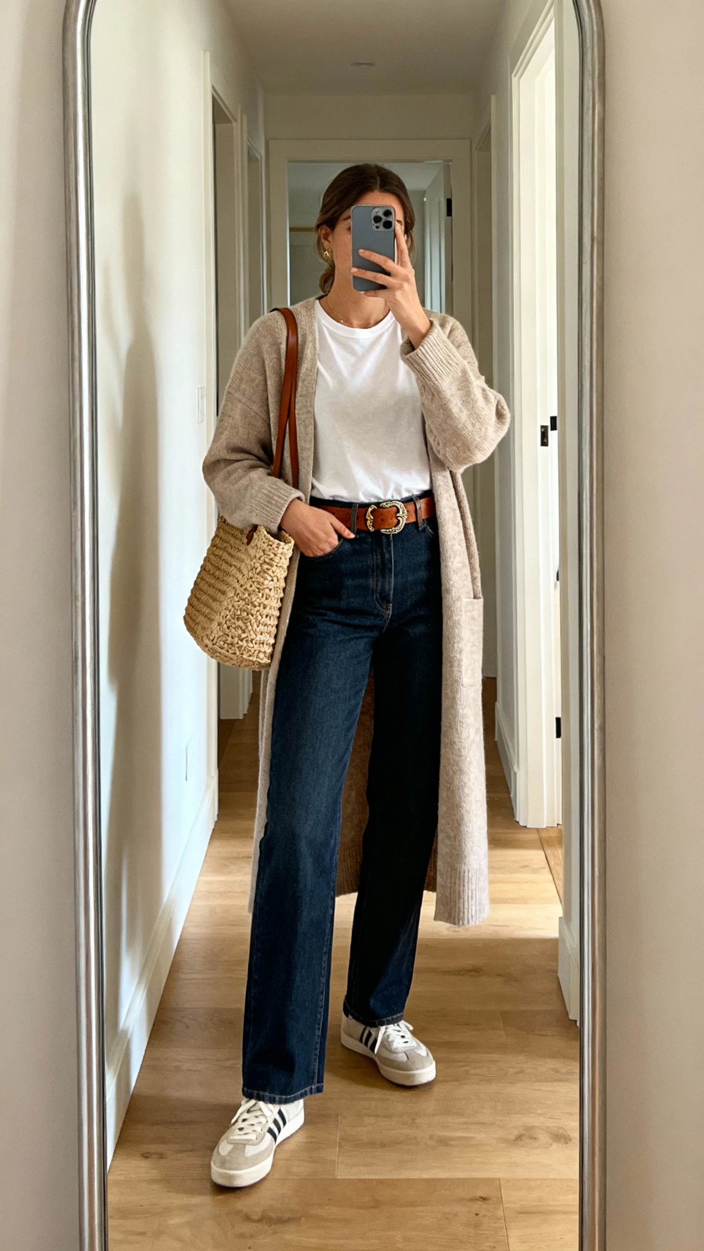 iPhone mirror selfie of a woman wearing a longline cardigan over a tee, tailored straight-leg jeans, artisan leather belt, and simple sneakers with a woven tote, face not visible, entryway full-length mirror, natural morning light, casual iPhone photo.