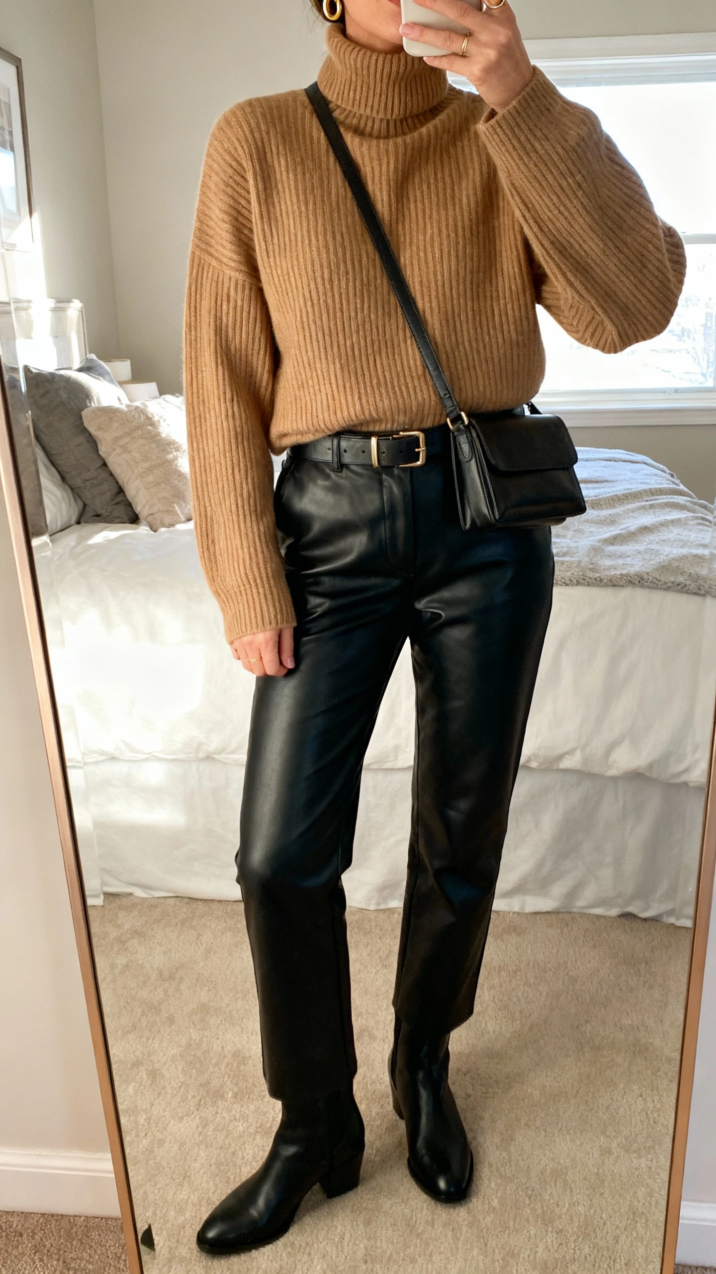 iPhone mirror selfie of a woman wearing a luxe ribbed cashmere turtleneck sweater in camel tucked into black leather straight-leg pants, sleek black ankle boots, slim leather belt, small gold hoops, and a structured crossbody, face not visible, cropped at neck, bedroom full-length mirror with morning window light, natural iPhone photo quality.