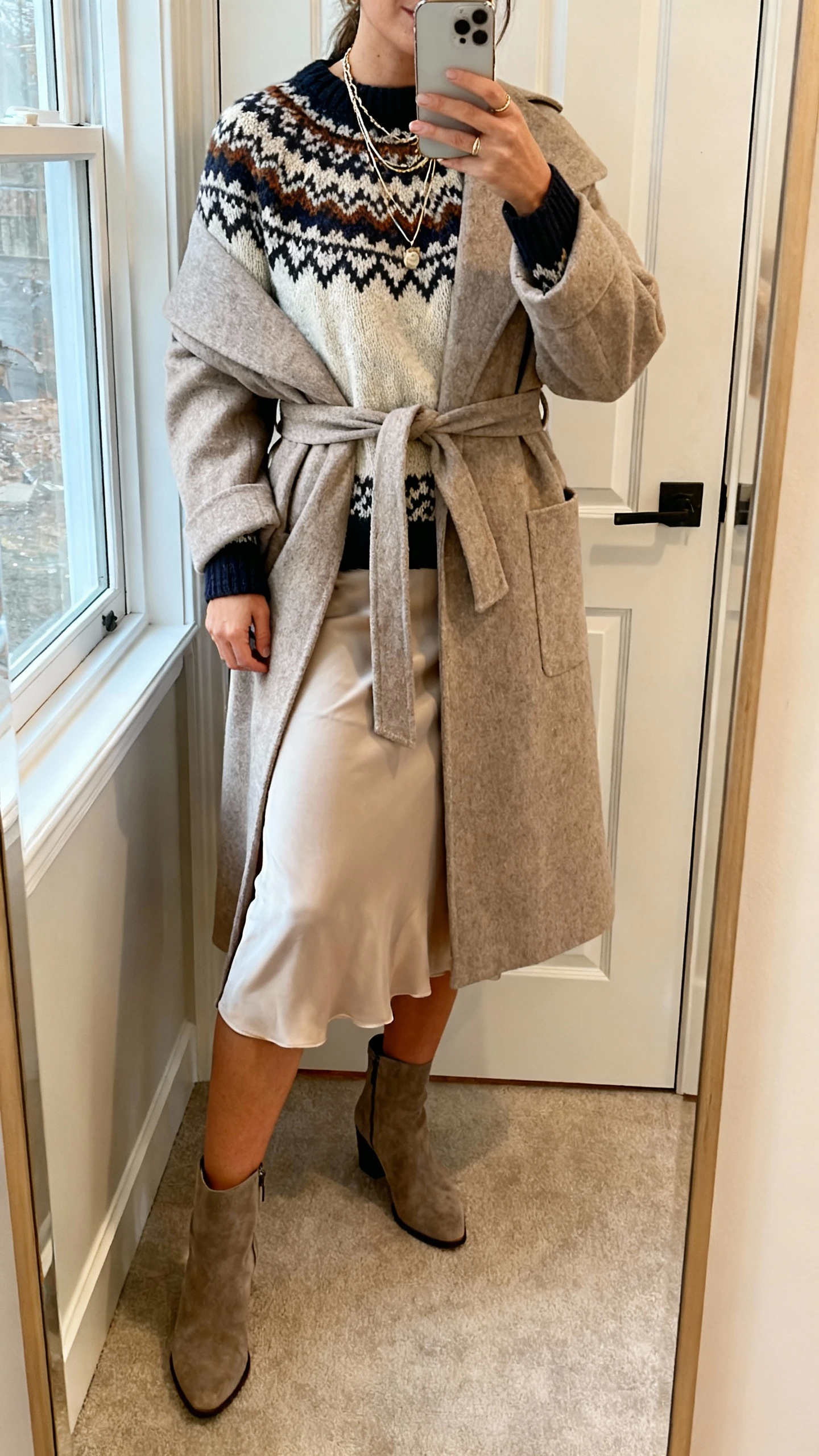 iPhone mirror selfie of a woman wearing a patterned knit sweater with a slip skirt and a belted wrap coat, layered necklaces and ankle boots, face not visible, entryway mirror by the door, natural window light, natural iPhone photo quality.