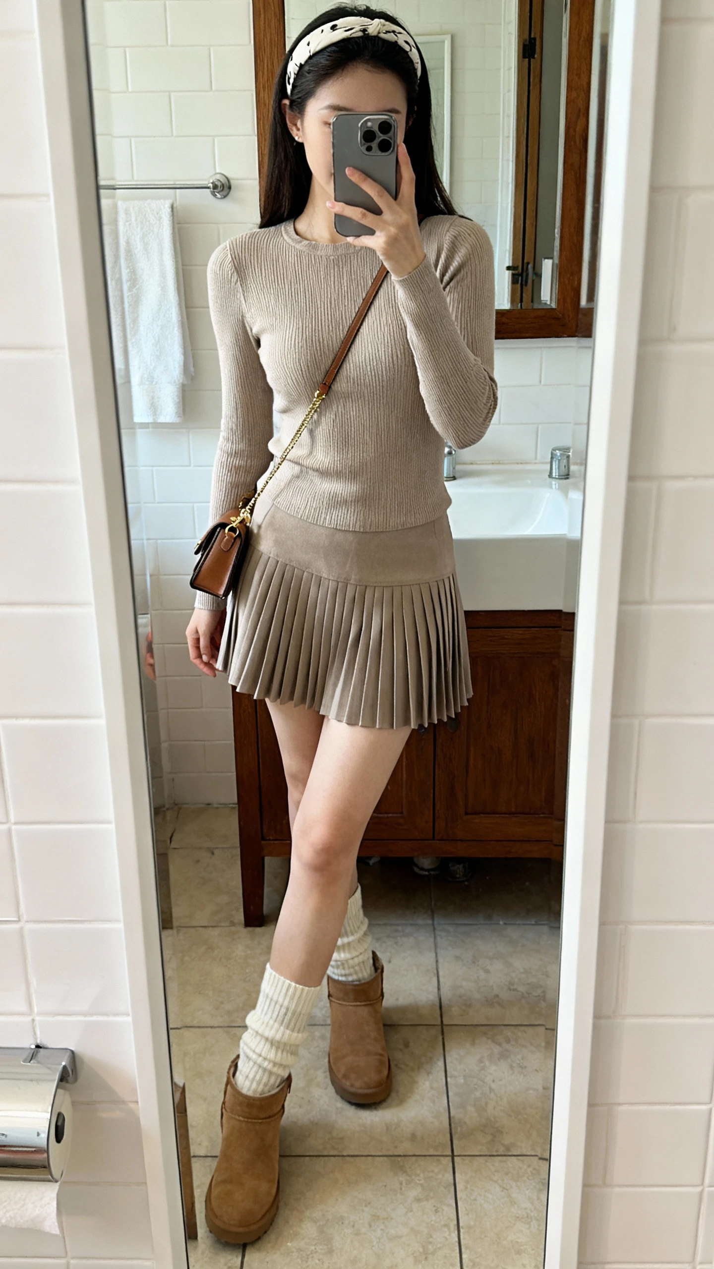 iPhone mirror selfie of a woman wearing a pleated mini skirt, fitted knit top, sock boots, headband, small shoulder bag, face not visible, cropped at neck, bathroom full-length mirror, soft indoor lighting, natural iPhone photo quality.