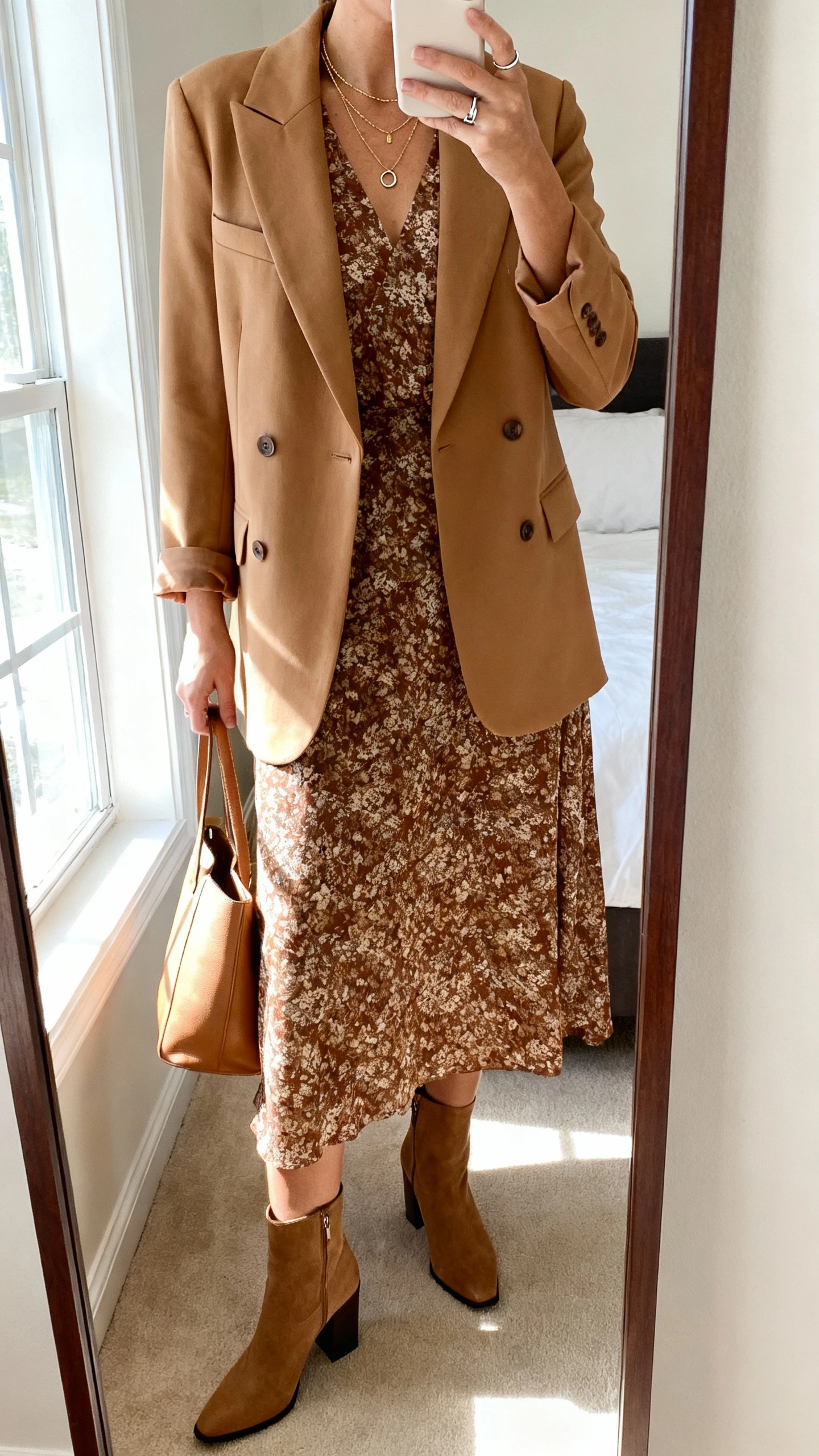 iPhone mirror selfie of a woman wearing a printed earthy-tone midi dress with a camel single-breasted blazer, block-heel ankle boots, layered delicate necklaces, a simple signet ring, and a minimal leather tote, face not visible, cropped at neck, bedroom mirror with window light, natural iPhone photo quality.