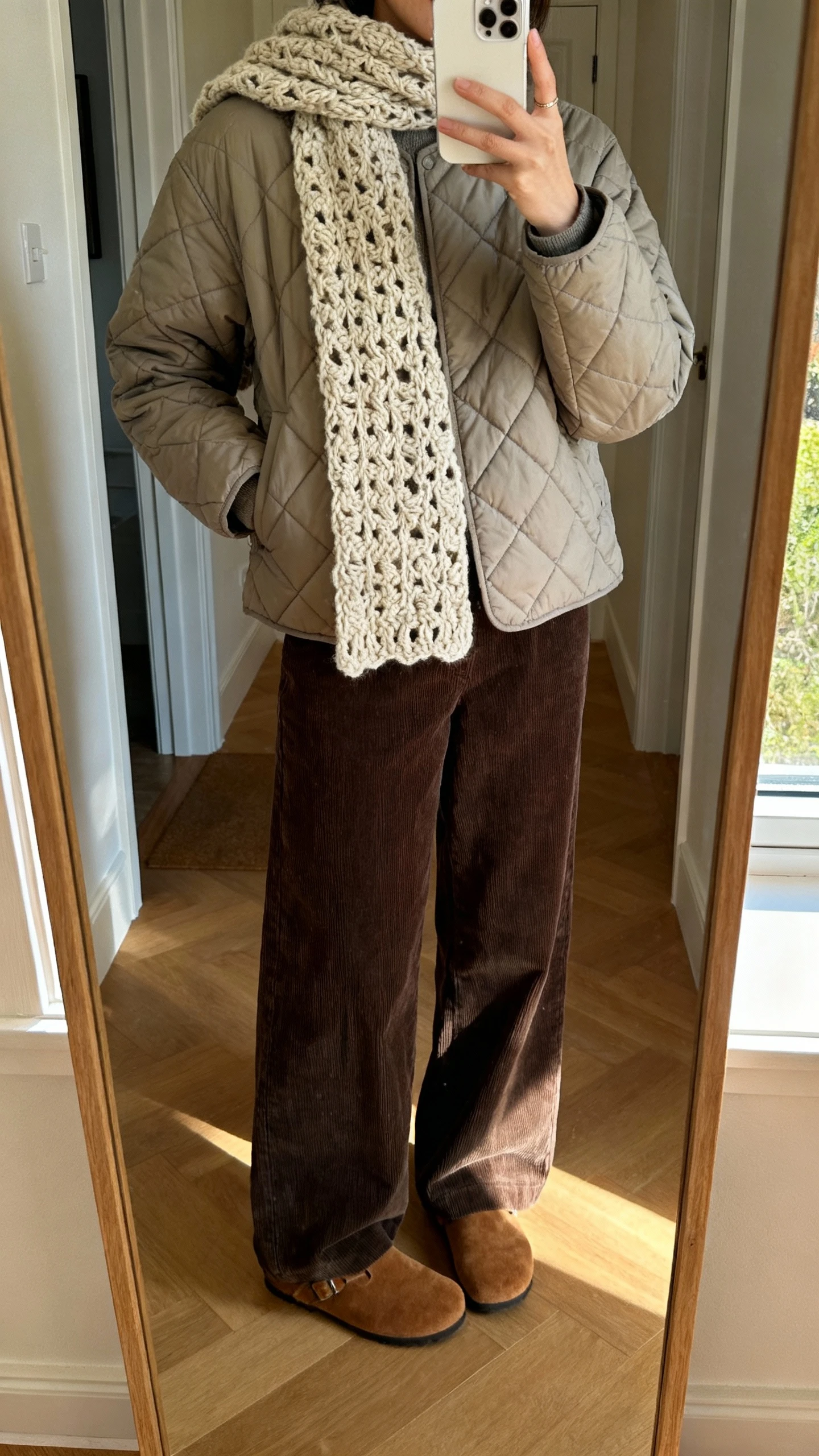 iPhone mirror selfie of a woman wearing a quilted jacket, wide-leg corduroy pants, and a chunky crochet scarf with suede clogs, face not visible, entryway mirror, soft afternoon window light, natural iPhone photo.