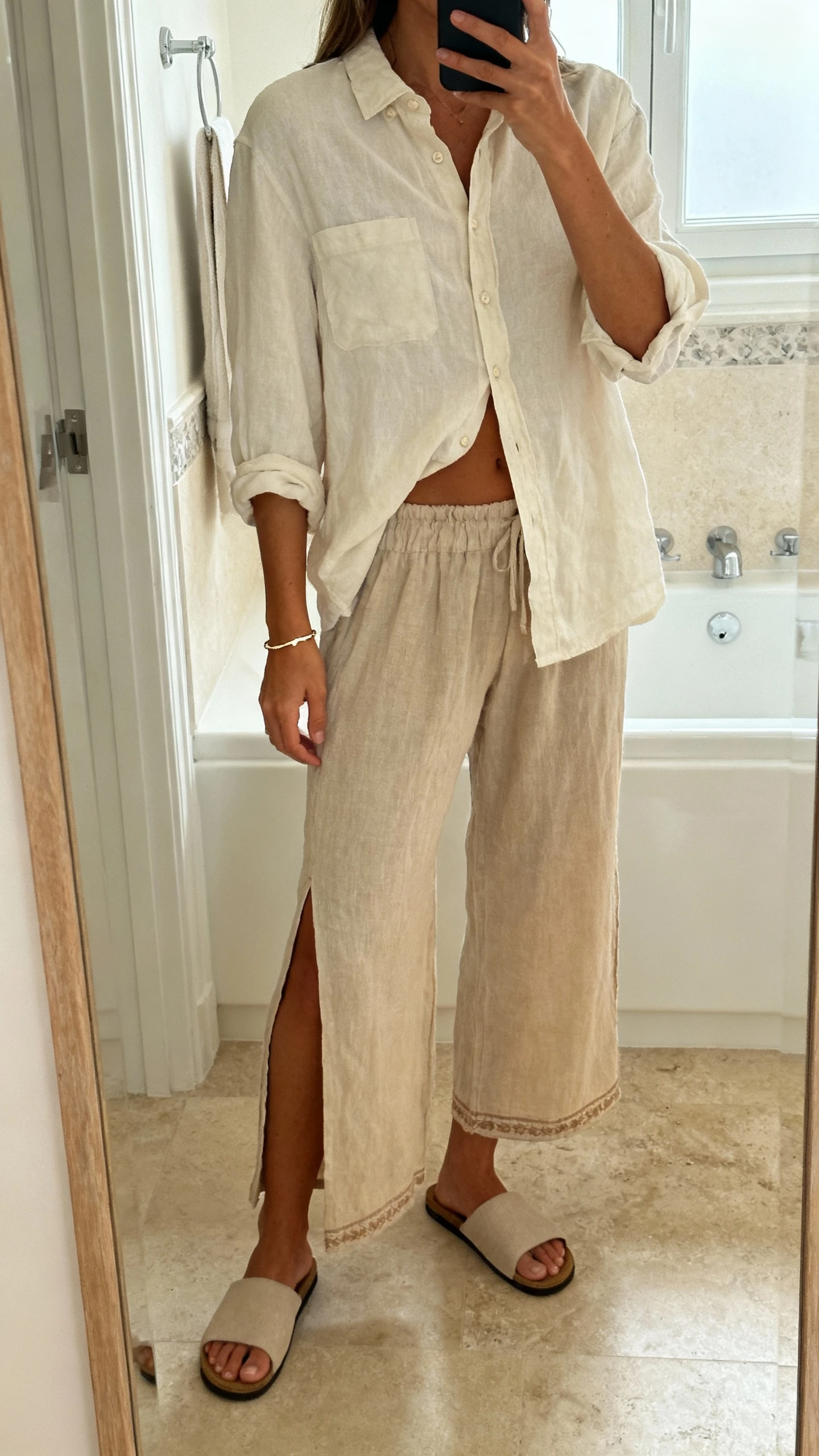 iPhone mirror selfie of a woman wearing a relaxed button-down shirt half-tucked into slit boho pants, delicate bracelet, minimalist slides, face not visible, bathroom mirror, soft indoor lighting, natural iPhone photo quality.