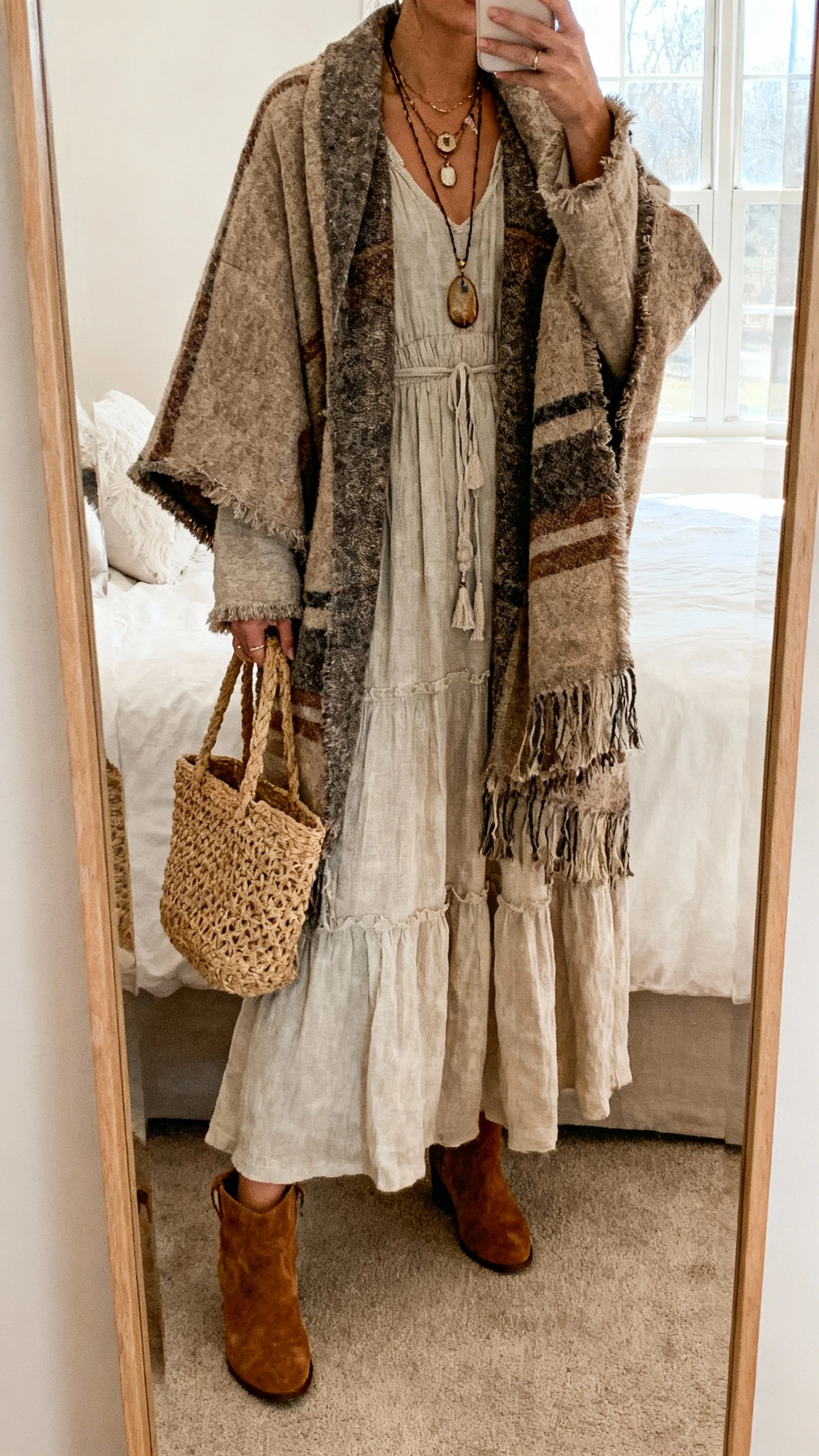 iPhone mirror selfie of a woman wearing a rustic blanket coat over a flowy boho maxi dress with layered pendant necklaces and suede ankle boots, holding a woven tote, face not visible, bedroom full-length mirror, soft window light, natural iPhone photo quality.