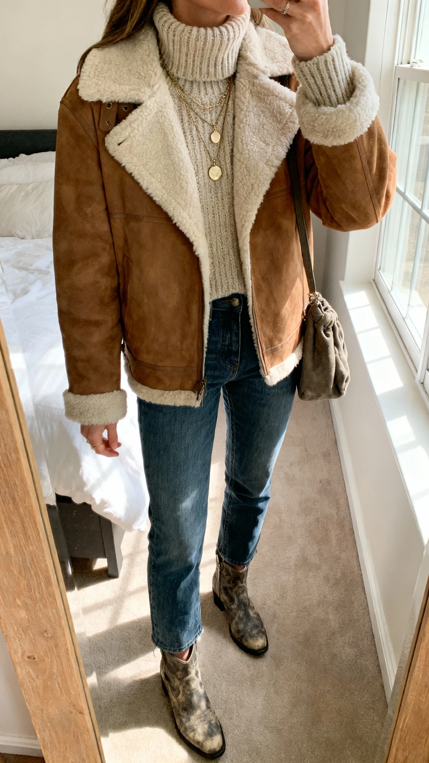 iPhone mirror selfie of a woman wearing a shearling-trimmed suede jacket over a chunky knit turtleneck, high-waisted straight jeans, rugged ankle boots, layered pendant necklaces, and a slouchy crossbody, face not visible, cropped at neck, bedroom mirror with soft morning window light, natural iPhone photo quality.