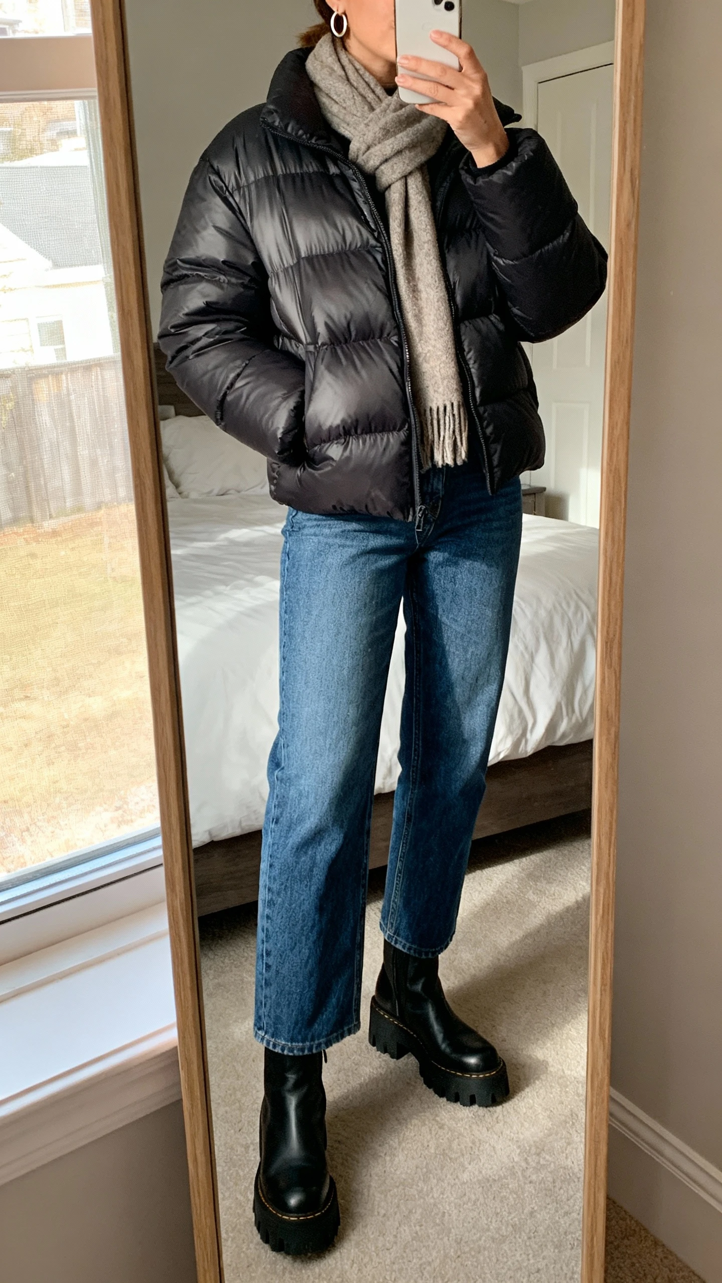 iPhone mirror selfie of a woman wearing a sleek elevated puffer jacket, straight-leg blue jeans, chunky-soled ankle boots, a cozy scarf, and minimal hoop earrings, face not visible, cropped at neck, bedroom full-length mirror with morning window light, natural iPhone photo quality.