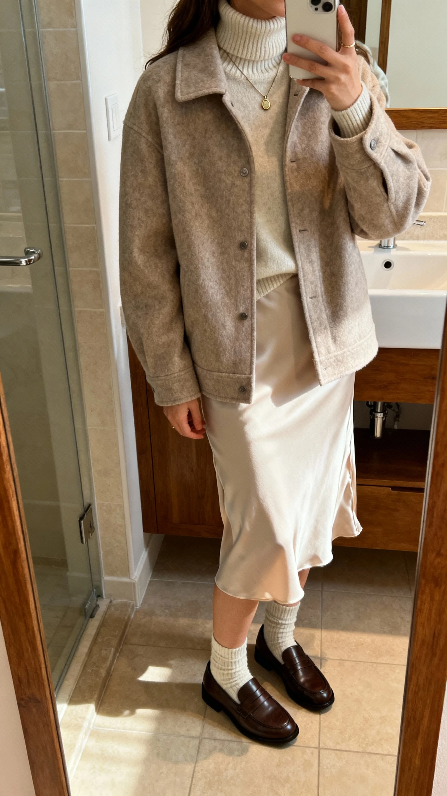 iPhone mirror selfie of a woman wearing a soft shacket layered over a slip skirt and a cozy turtleneck, with socks and loafers and a subtle pendant, face not visible, bathroom mirror, warm indoor light, natural iPhone photo quality.