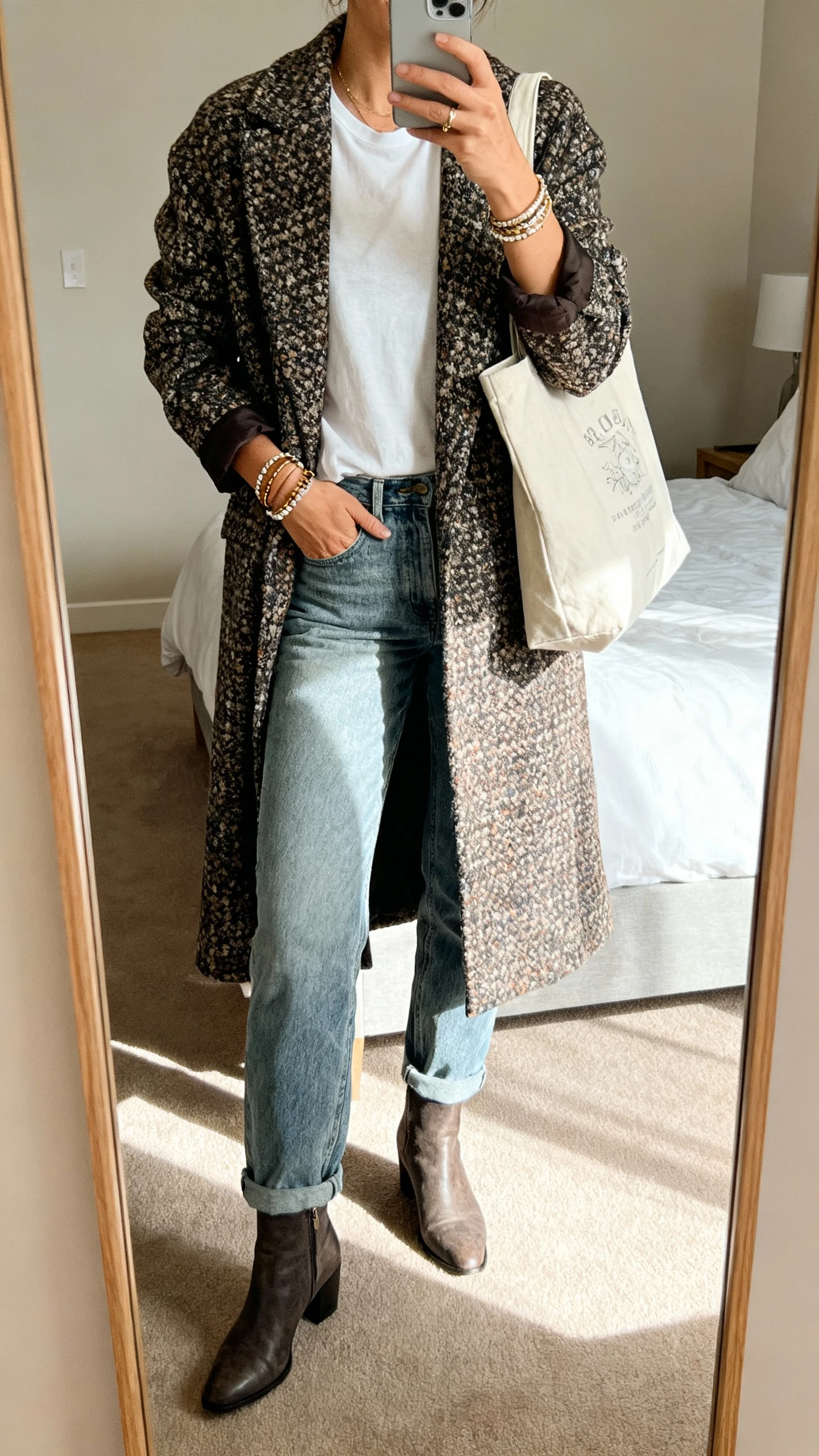 iPhone mirror selfie of a woman wearing a statement coat over boho basics (simple tee, relaxed jeans, ankle boots) with layered bracelets and a tote bag, face not visible, full-length bedroom mirror, natural morning light, natural iPhone photo quality
