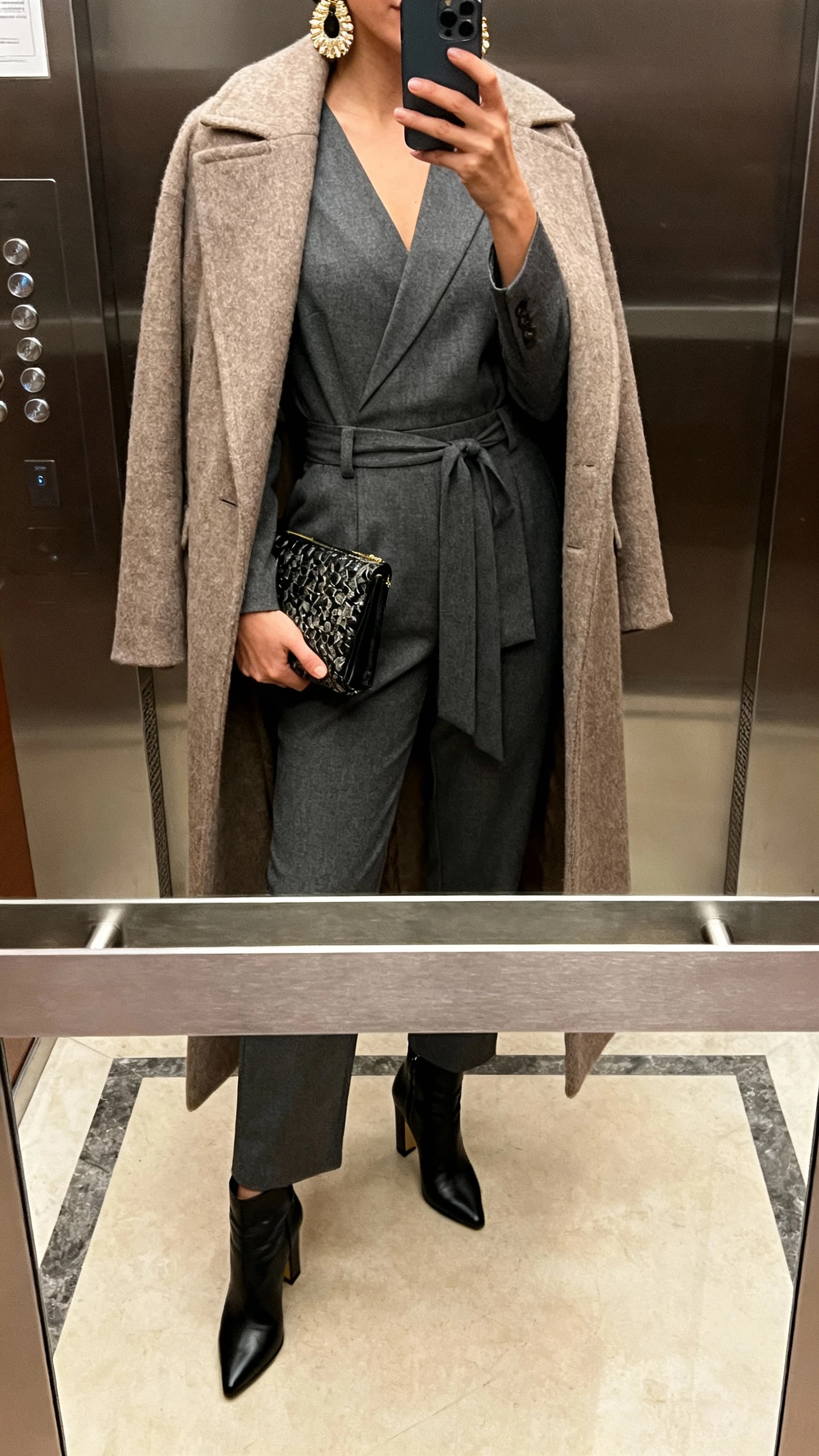 iPhone mirror selfie of a woman wearing a tailored jumpsuit with a long wool coat as winter armor, heeled booties, statement earrings, and a clutch, face not visible, cropped at neck in an elevator mirror, indoor lighting, natural iPhone photo quality