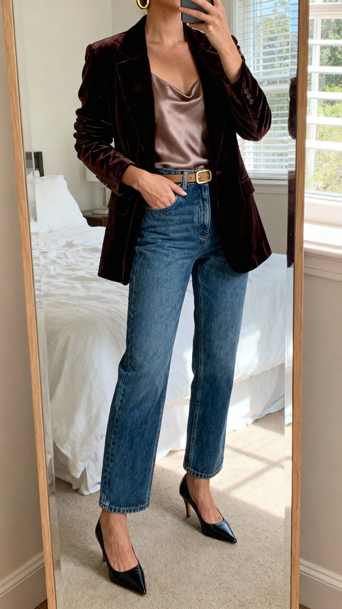iPhone mirror selfie of a woman wearing a velvet blazer over a silky camisole, elevated straight-leg denim, pointed-toe heels, gold hoops and a slim belt, face not visible, cropped at neck in a bedroom mirror, natural window light, natural iPhone photo quality
