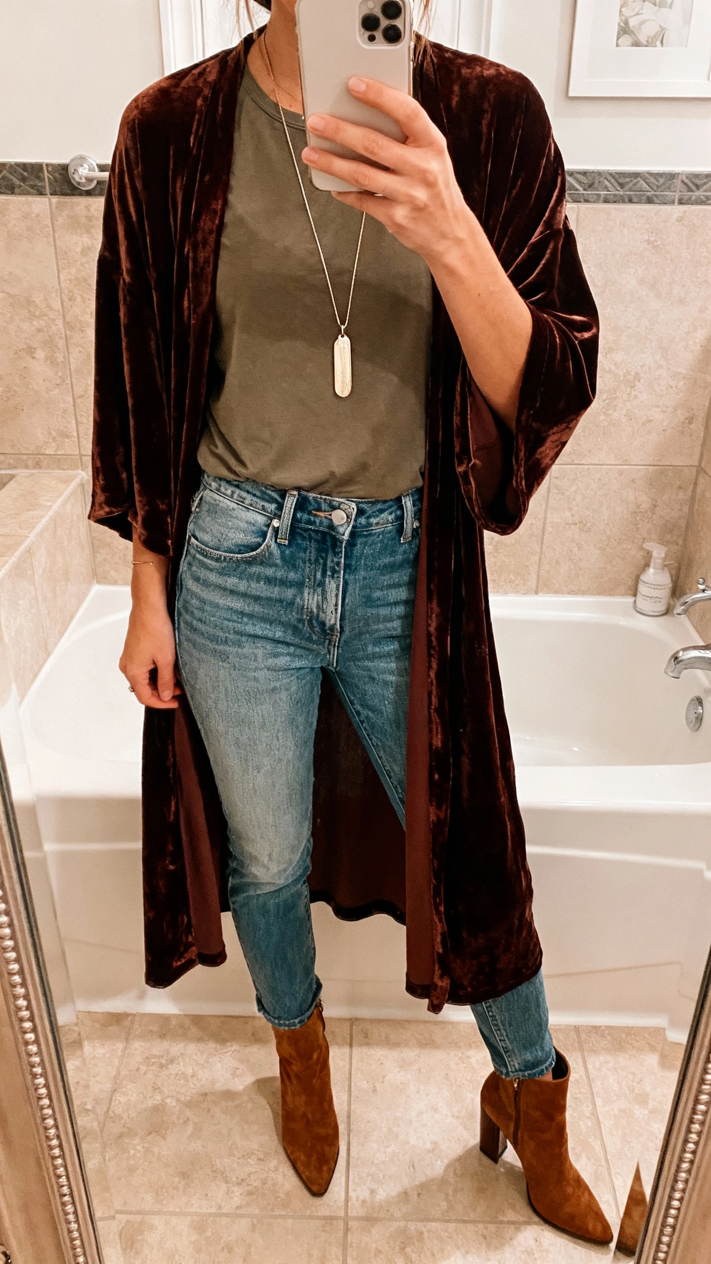 iPhone mirror selfie of a woman wearing a velvet duster over a tee and high-rise denim with heeled booties and a long pendant necklace, face not visible, bathroom mirror, warm indoor lighting, natural iPhone aesthetic