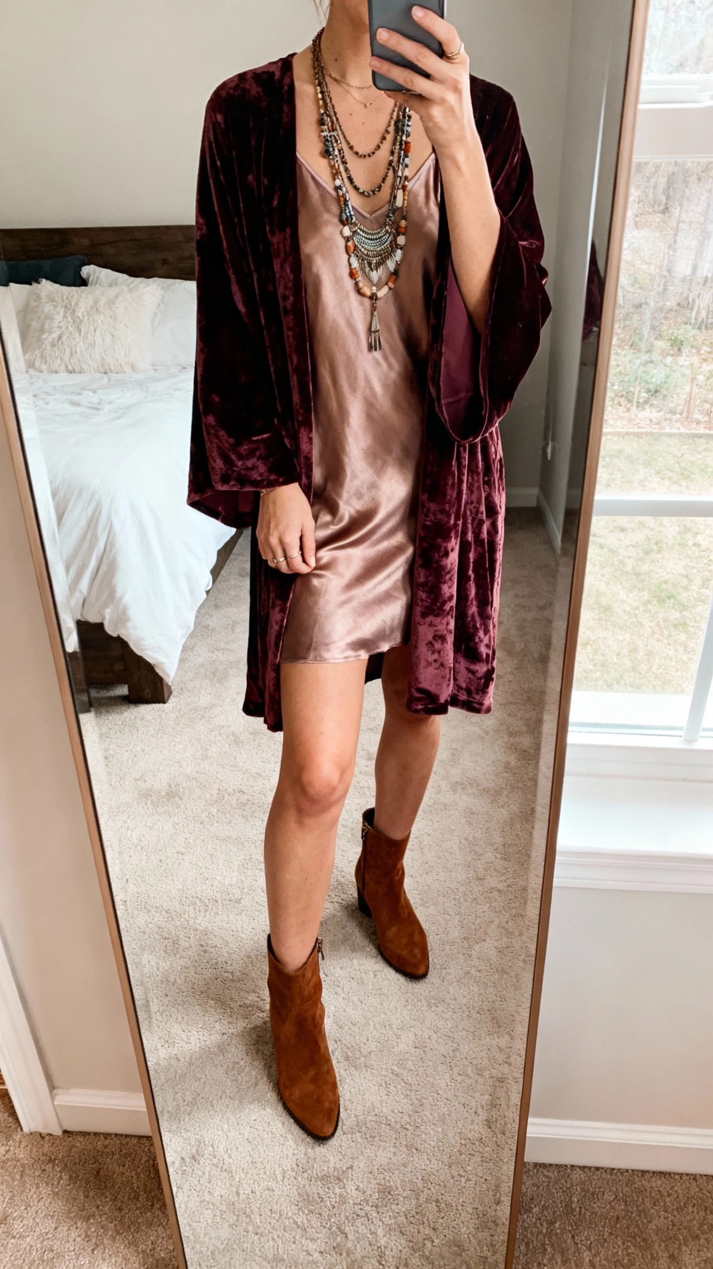 iPhone mirror selfie of a woman wearing a velvet kimono over a satin slip dress with ankle boots and layered boho necklaces, face not visible, cropped at neck, bedroom full-length mirror, soft window light, natural iPhone photo quality.