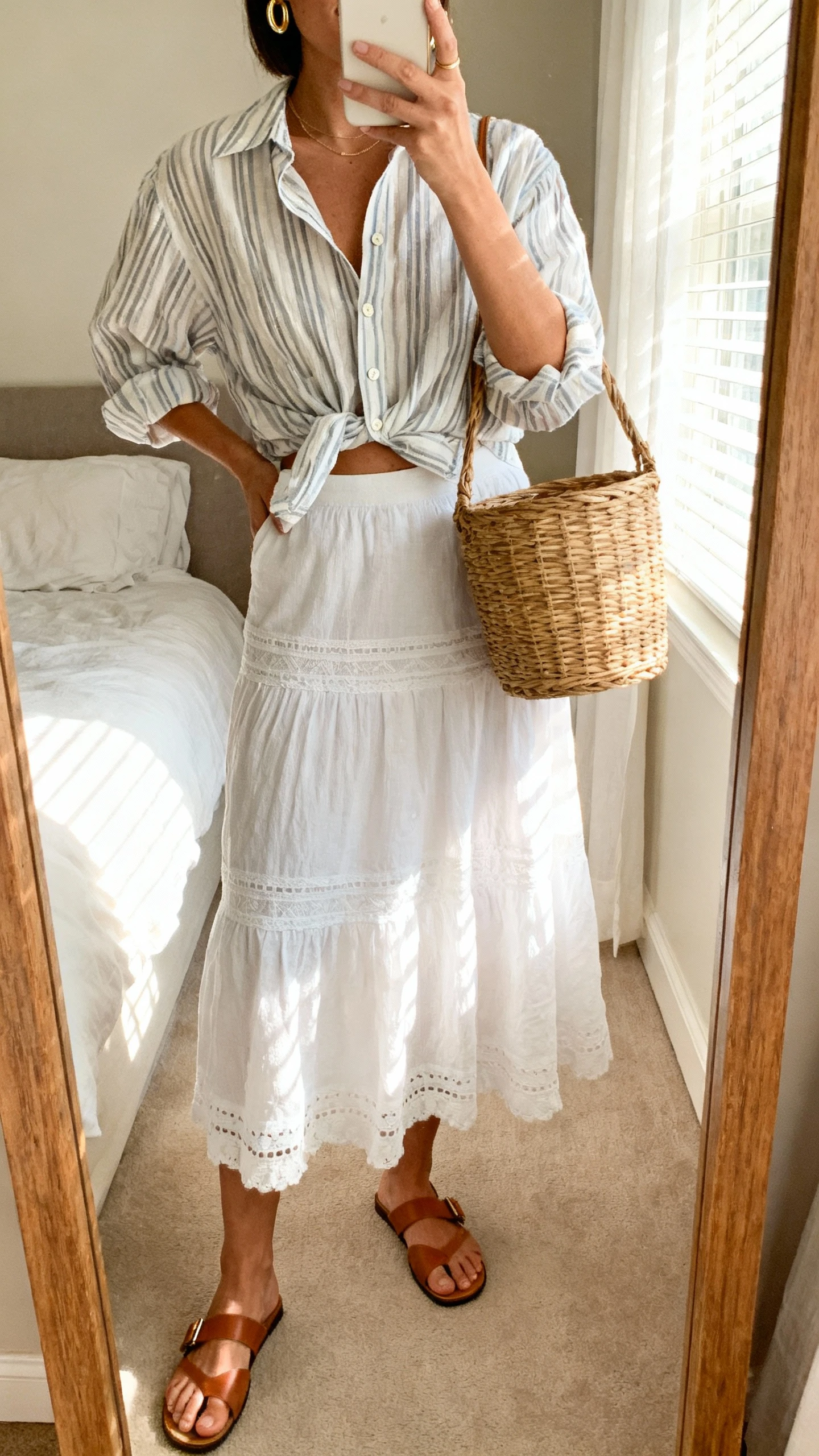 iPhone mirror selfie of a woman wearing a white boho skirt with a breezy striped button-up half-tucked, basket bag, flat leather sandals, delicate gold hoops, face not visible, cropped at neck, bedroom mirror with morning window light, natural iPhone photo quality.