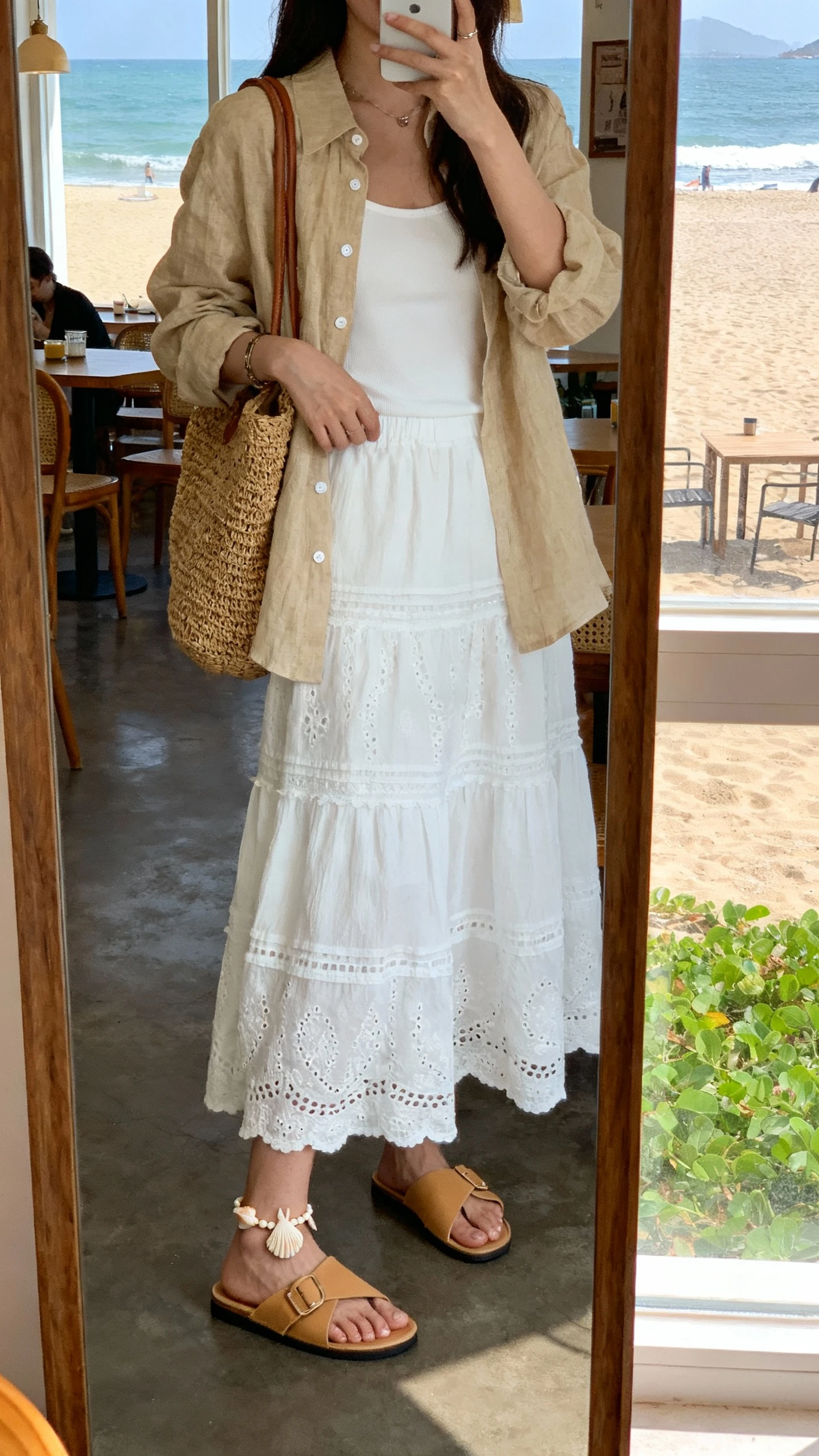 iPhone mirror selfie of a woman wearing a white boho skirt with a sand linen button-up layered over a white camisole, tan slide sandals, woven tote, shell anklet, face not visible, beachside café mirror, bright window light, iPhone photo quality.