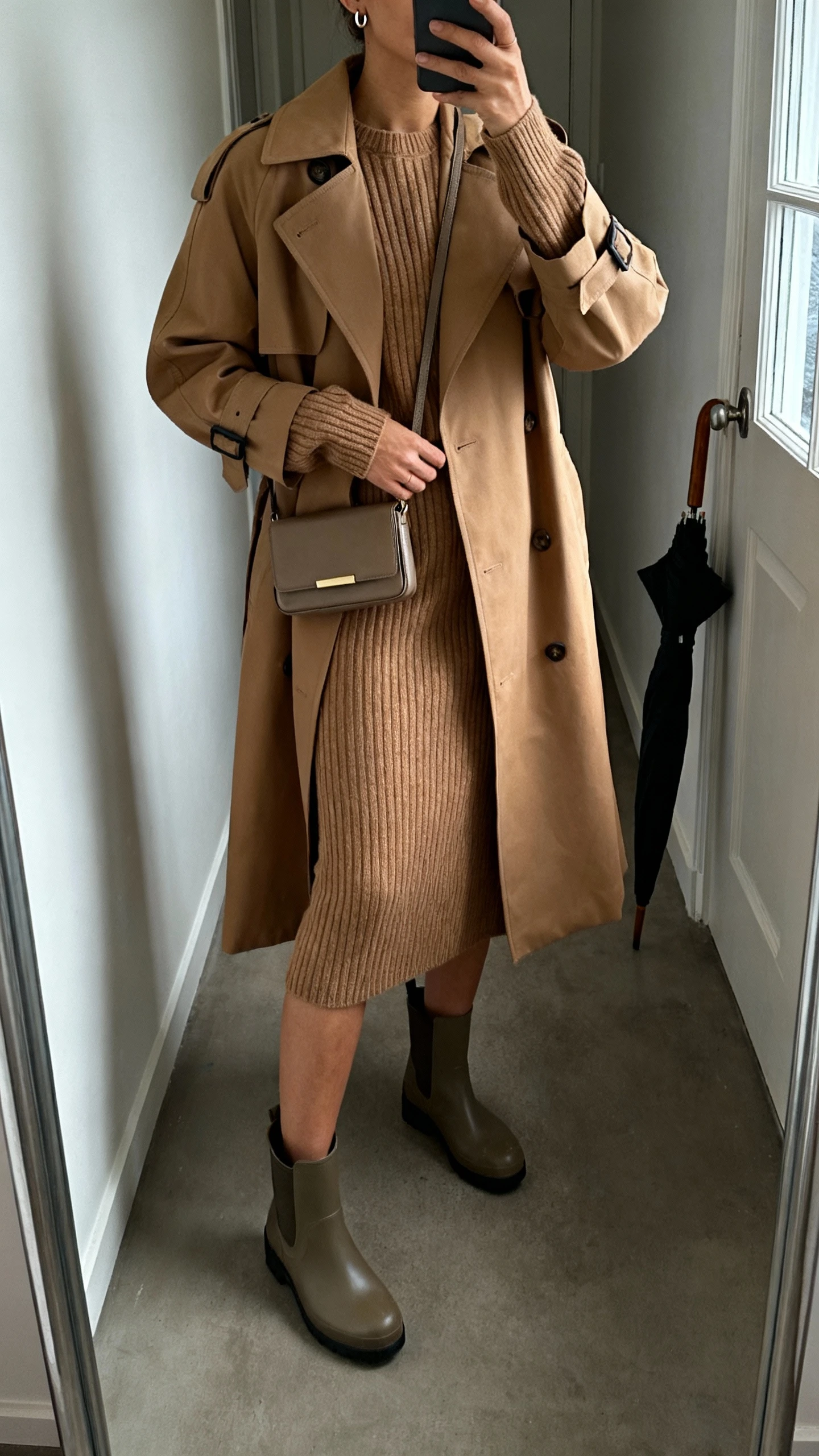 iPhone mirror selfie of a woman wearing an elevated camel trench over a matching ribbed knit set (sweater and skirt), waterproof ankle boots, compact crossbody, tiny hoops; face not visible, cropped at neck, hallway mirror with umbrella by the door, overcast window light, natural iPhone photo quality.