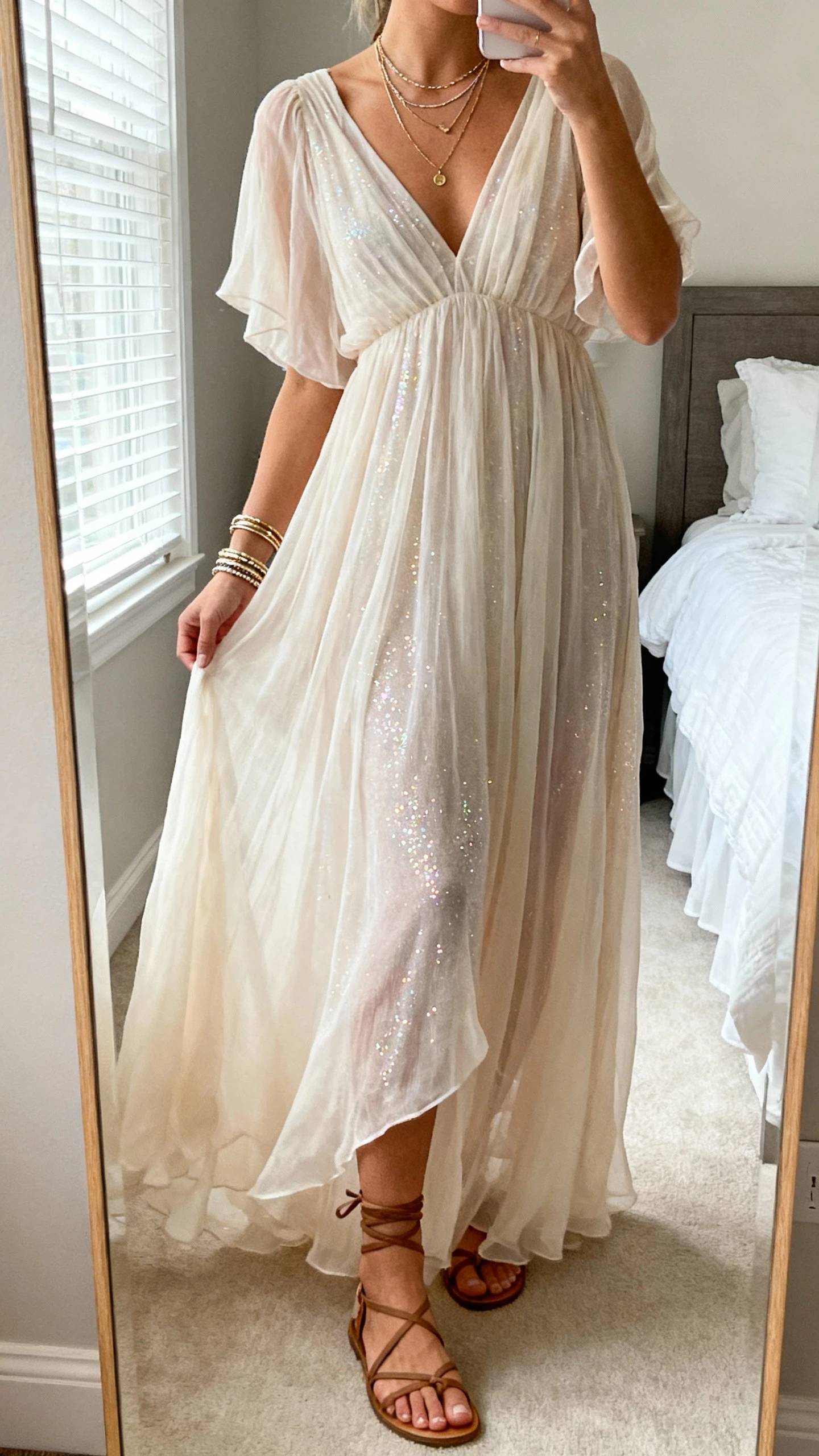 iPhone mirror selfie of a woman wearing an ethereal flowy maxi dress with a hint of sparkle, strappy sandals, delicate layered necklaces and bangles, face not visible, cropped at neck, bedroom mirror with soft window light, natural iPhone photo quality.