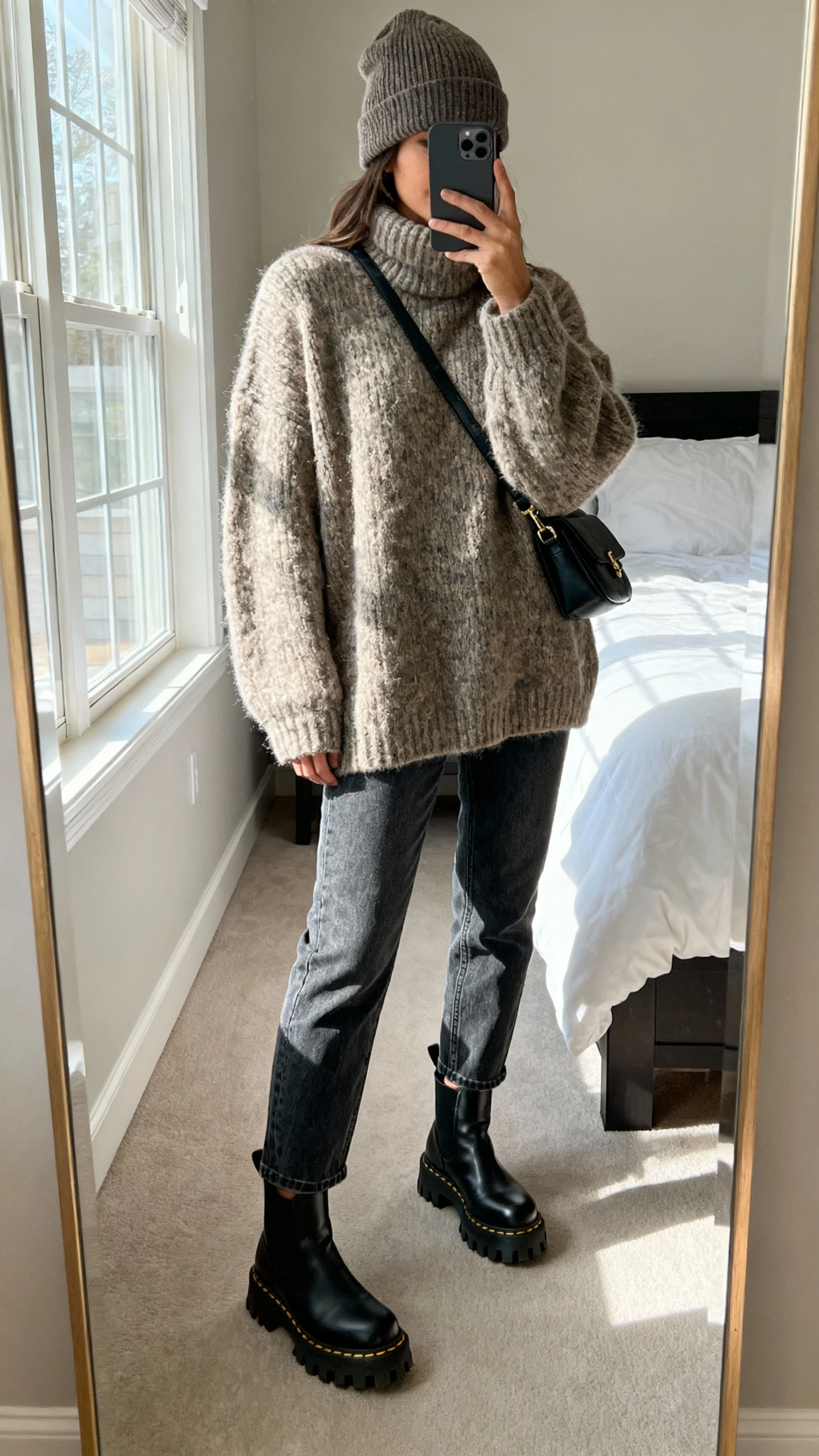 iPhone mirror selfie of a woman wearing an oversized chunky sweater, straight-leg jeans, lug-sole ankle boots, slouchy beanie, crossbody bag, face not visible, cropped at neck, bedroom mirror with natural window light, natural iPhone photo quality.