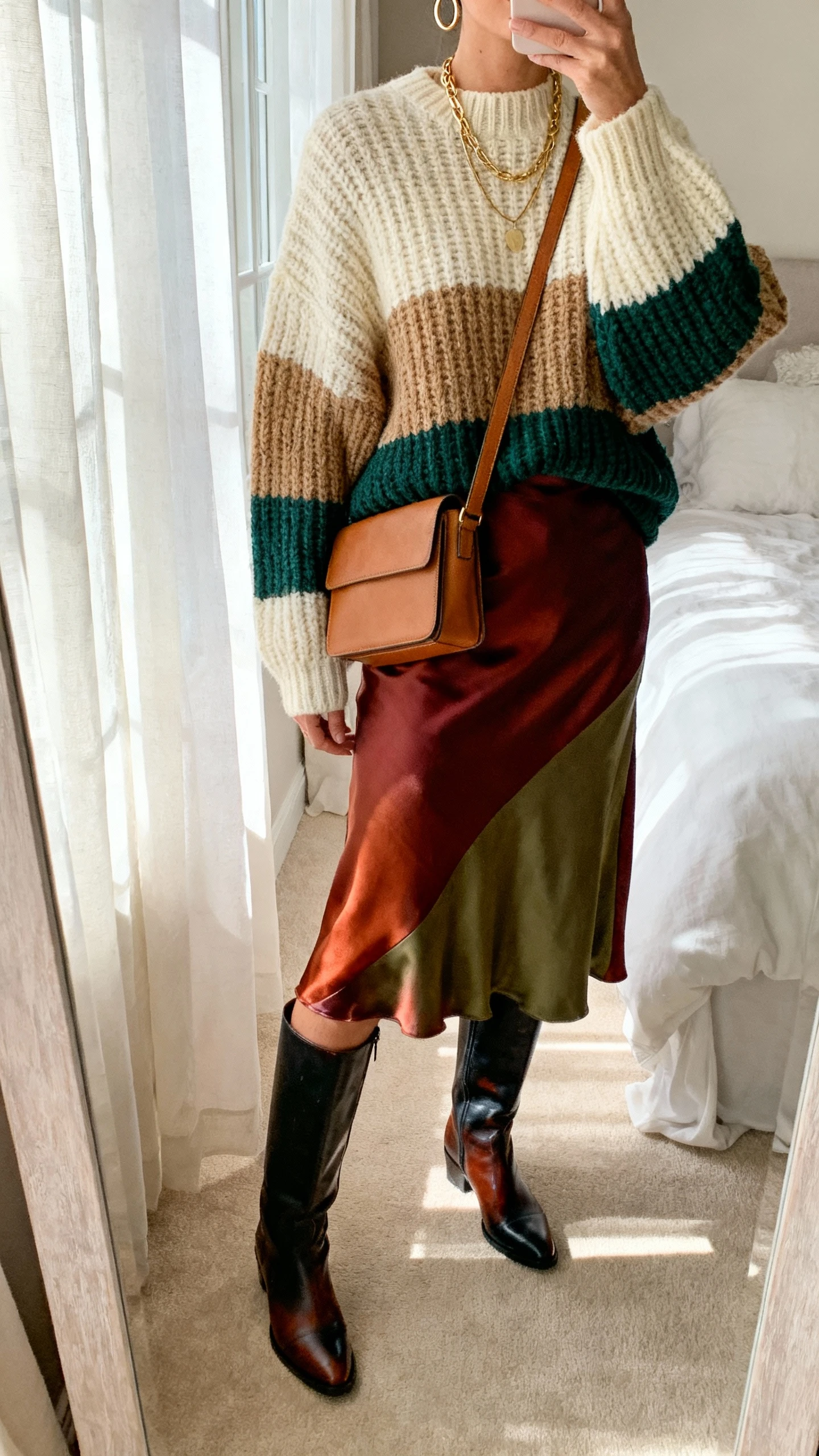 iPhone mirror selfie of a woman wearing an oversized chunky knit sweater (cream/camel/forest green), satin or chiffon midi skirt (rust/burgundy/olive), knee-high leather boots (brown/black), structured tan crossbody, layered gold necklaces and simple hoops, face not visible, cropped at neck in a bedroom mirror with window light, natural iPhone photo quality.