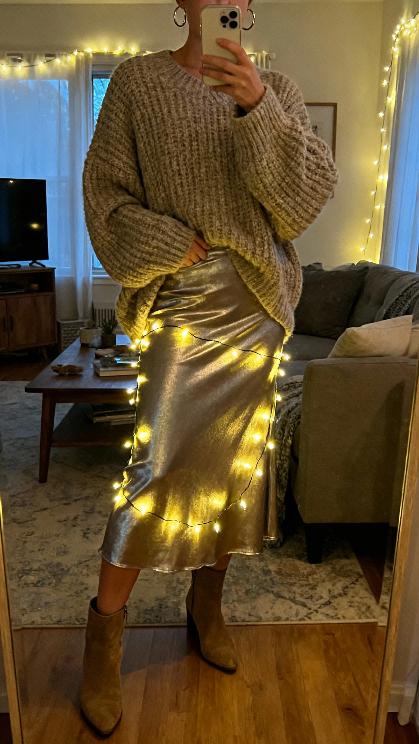 iPhone mirror selfie of a woman wearing an oversized chunky cardigan, metallic midi skirt that glows under string lights, ankle boots, simple hoop earrings, face not visible, cozy living room with twinkle lights, evening indoor lighting, natural iPhone photo quality.