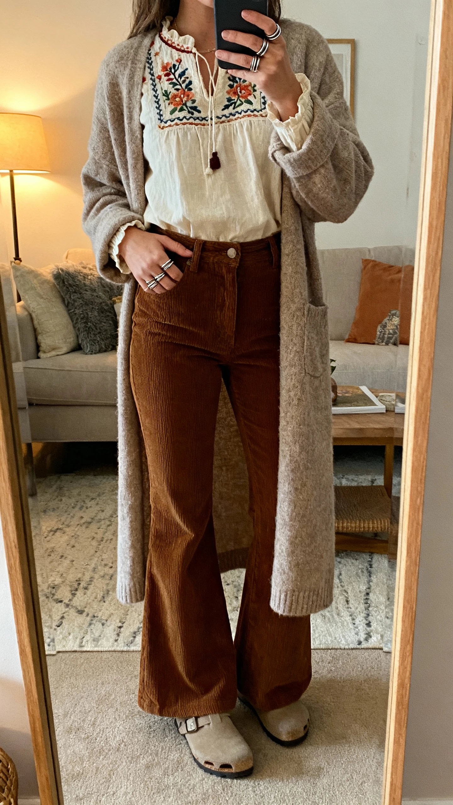 iPhone mirror selfie of a woman wearing flared corduroys, an embroidered blouse, and a longline cardigan with stacked rings and clogs, face not visible, cozy living room mirror, warm indoor lamp light, natural iPhone photo.