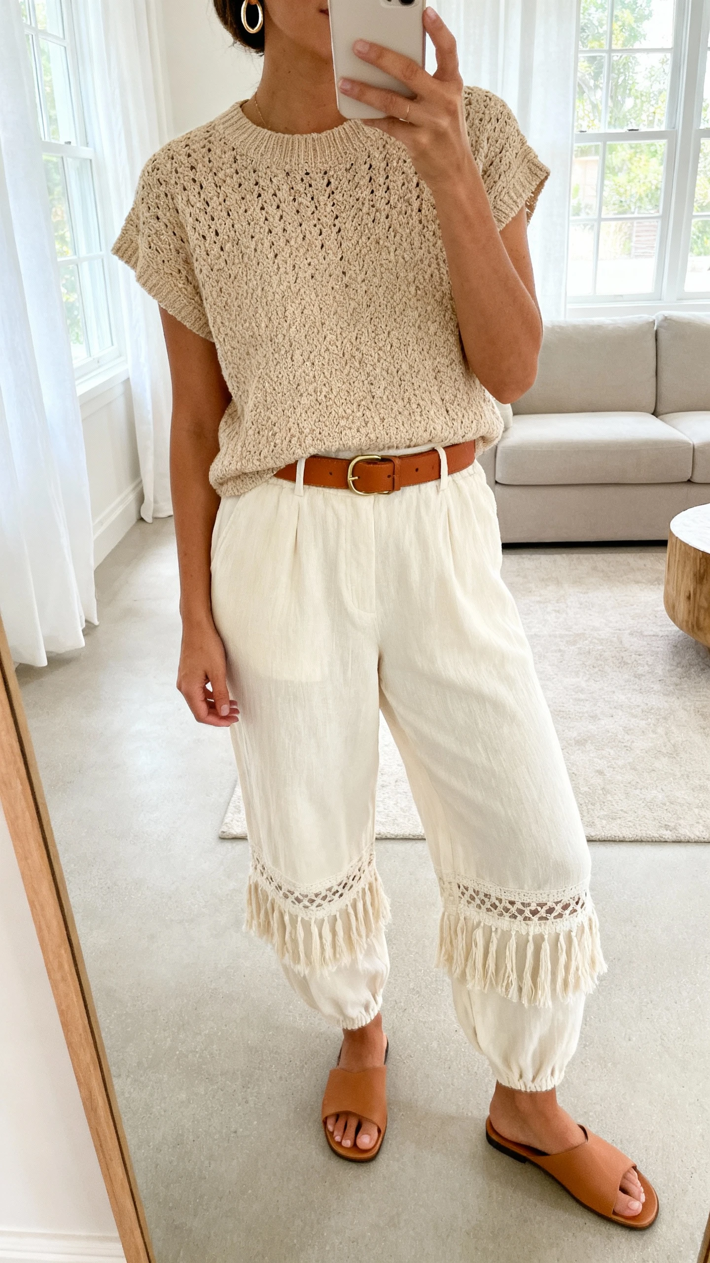 iPhone mirror selfie of a woman wearing head-to-toe neutrals: beige textured knit top with cream boho pants, tan belt, camel slides, subtle hoop earrings, face not visible, minimal living room mirror, soft window light, natural iPhone photo quality.
