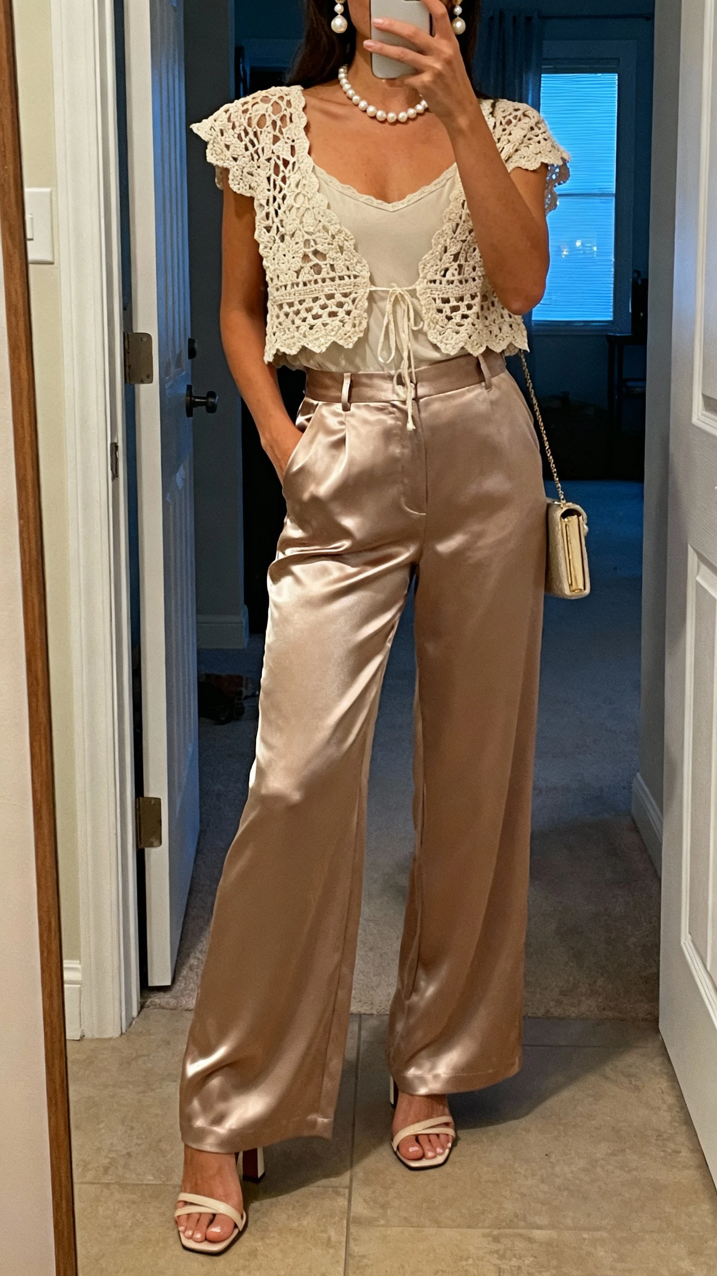 iPhone mirror selfie of a woman wearing satin wide-leg trousers with a crochet top layered over a camisole, pearl necklace and earrings, heeled sandals, and a small clutch, face not visible, entryway mirror with soft evening indoor light, natural iPhone photo quality