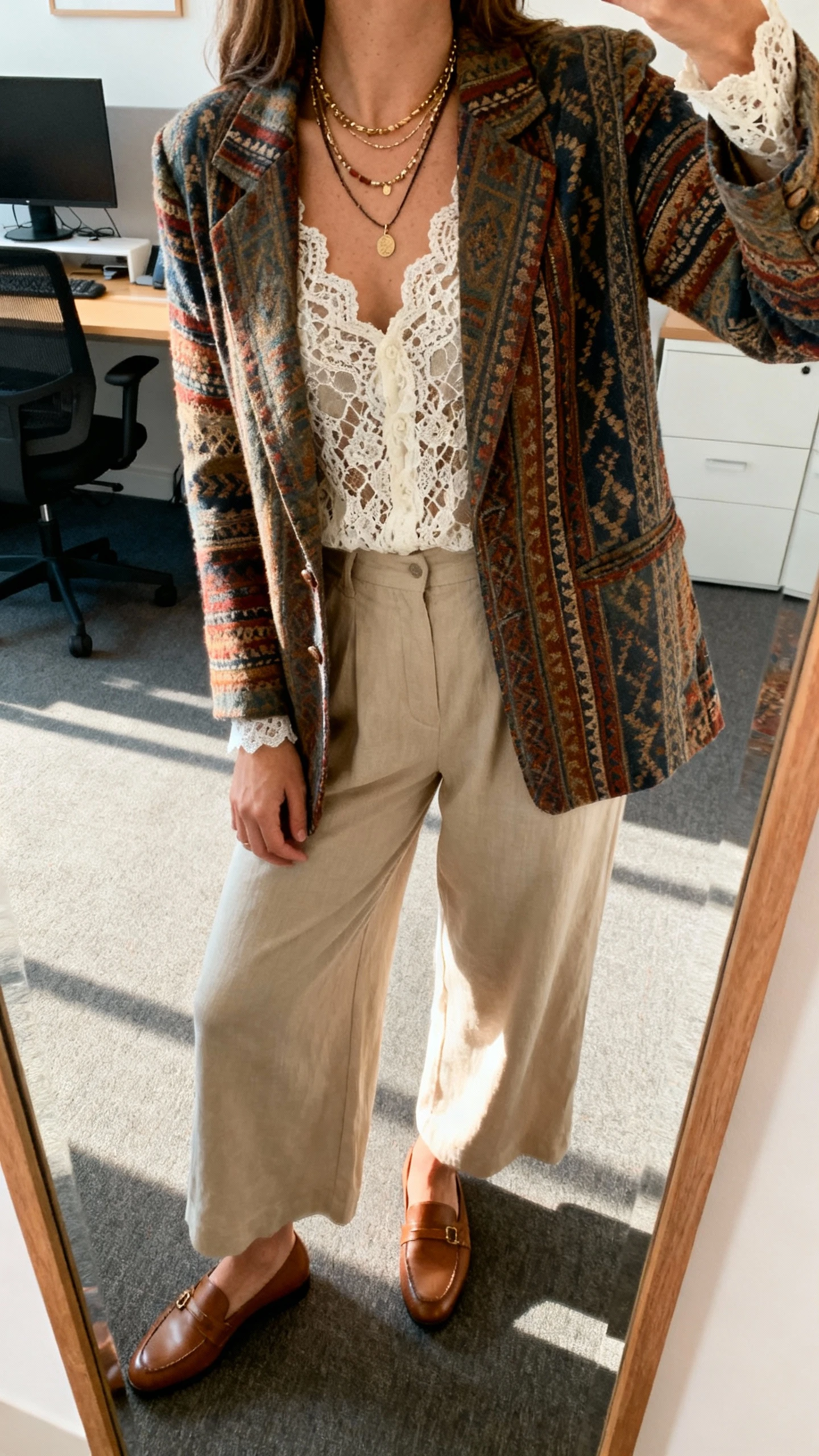 iPhone office mirror selfie of a woman in a boho blazer, wide-leg trousers, and a lace blouse, layered necklaces and loafers, face not visible, cropped at neck, afternoon indoor light, natural iPhone photo quality.
