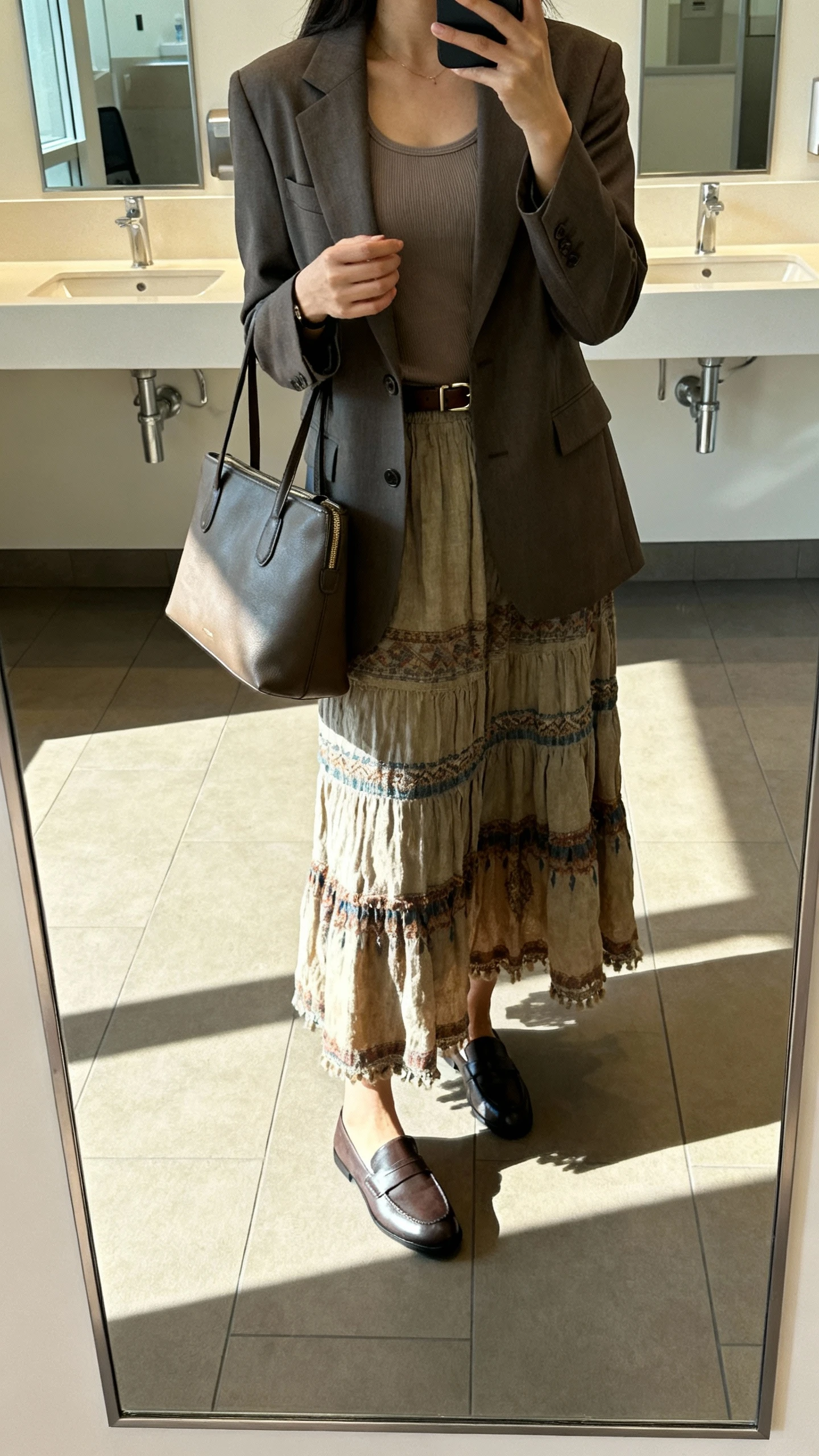 iPhone office mirror selfie of a woman wearing a long boho skirt with a tailored blazer over a fitted tank, sleek loafers, structured tote, subtle belt, face not visible, cropped at shoulders, corporate restroom mirror, bright morning light, natural iPhone aesthetic