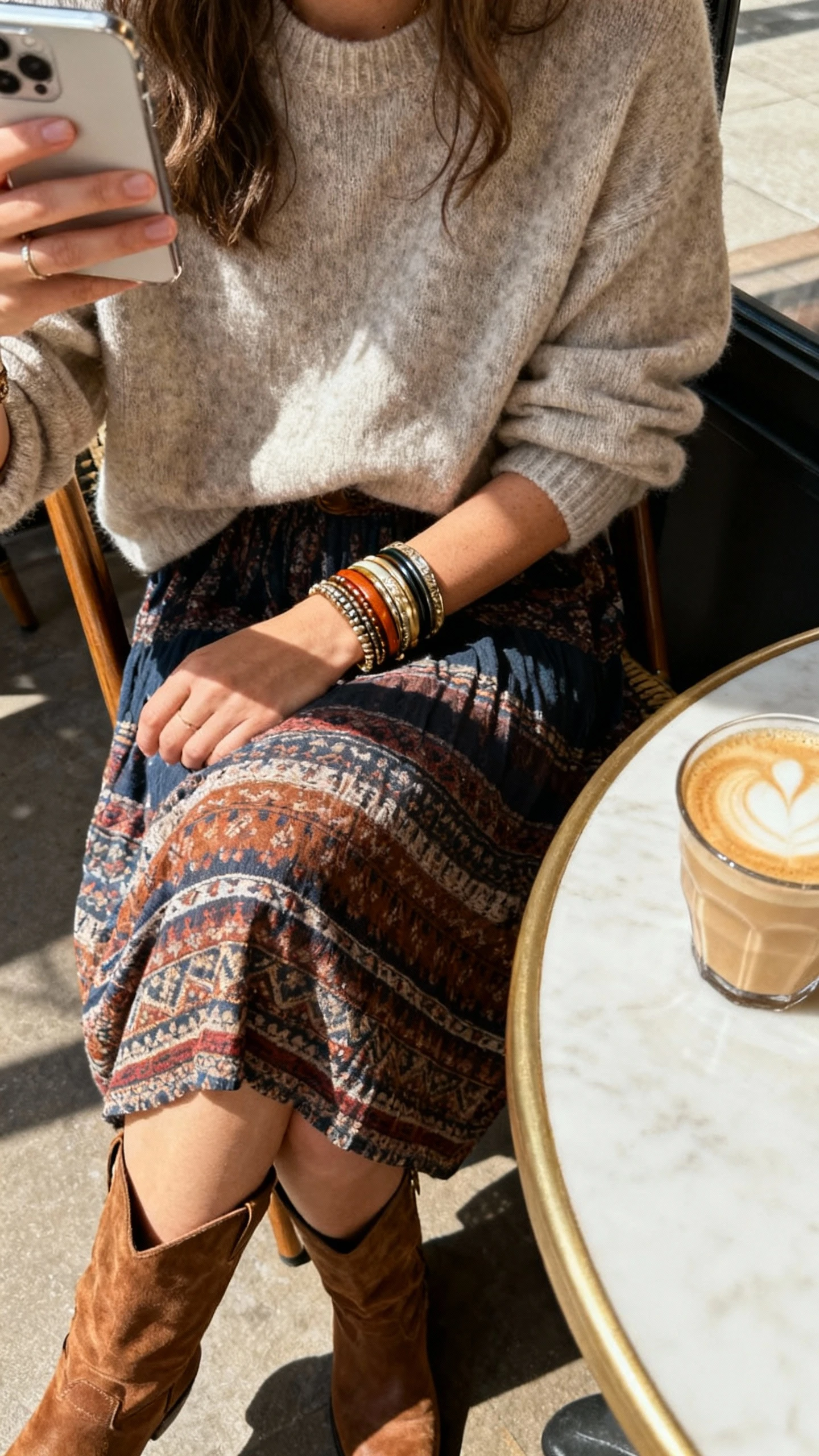 iPhone seated cafe selfie cropped at shoulders showing a woman wearing a relaxed sweater layered over a boho midi skirt with knee boots, stack of bangles, latte on table, face not visible, bright morning light, natural iPhone photo.