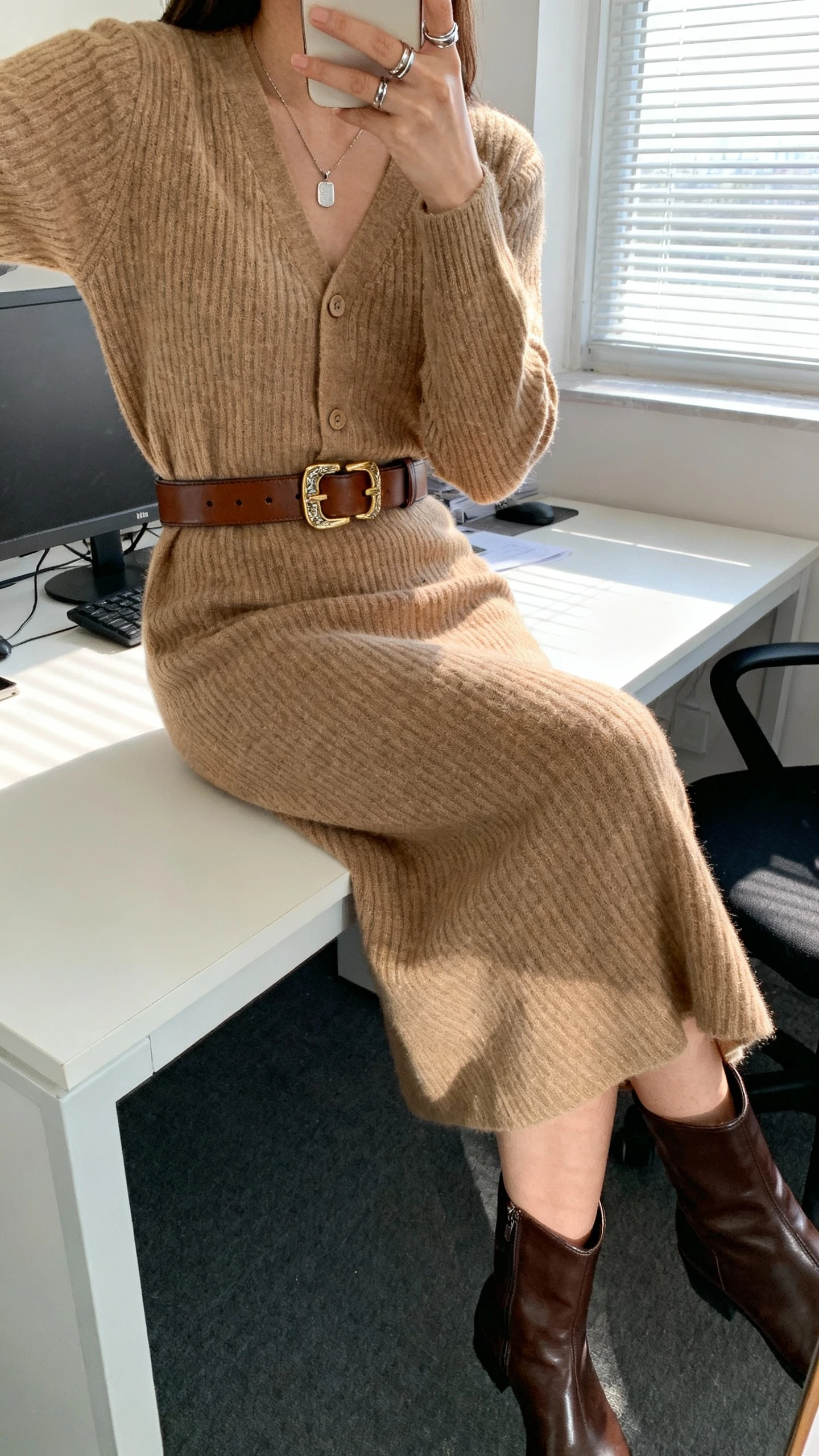 iPhone seated desk selfie of a woman wearing a tan fine-knit cardigan with matching midi knit skirt, a wide leather belt with a distinctive buckle, sleek leather boots, a simple pendant necklace, and stacked rings, face not visible, cropped at neck, office setting with window light, natural iPhone quality.