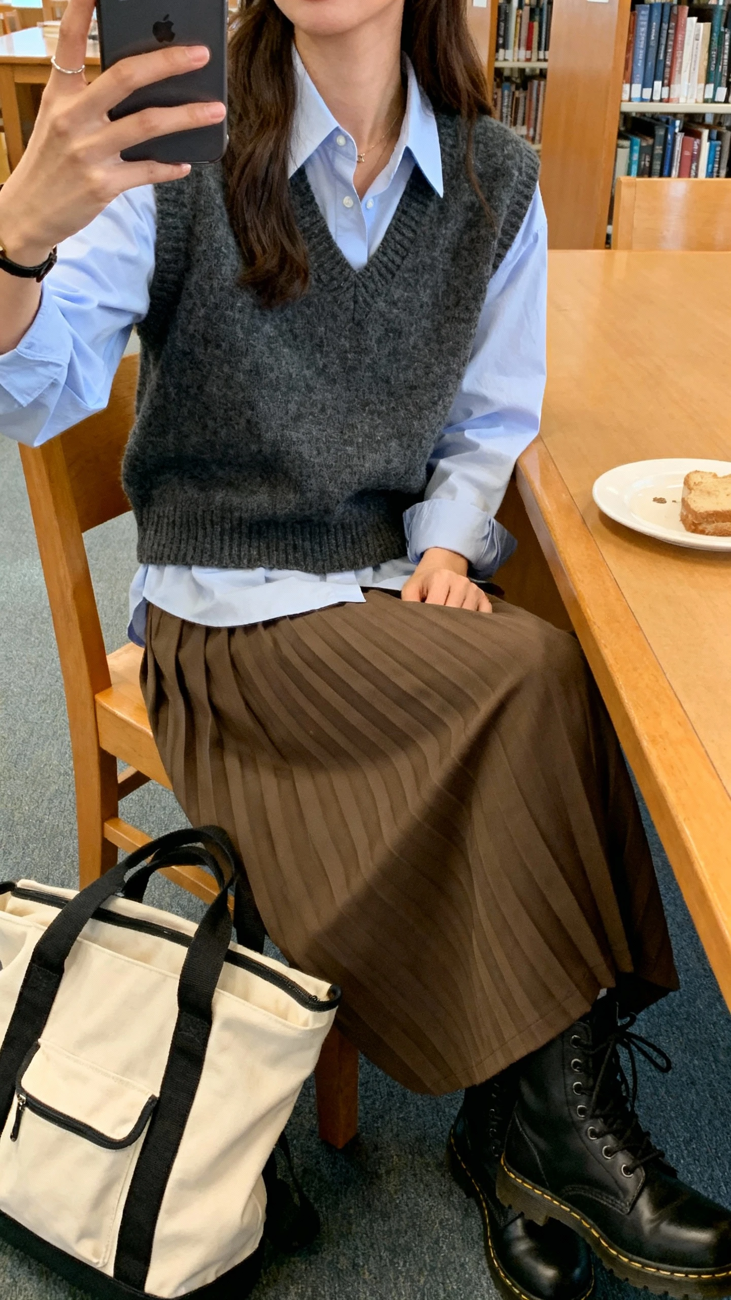 iPhone seated selfie cropped at neck showing a woman layering a sweater vest over an oxford shirt with pleated trousers, combat boots, tote backpack, library table setting, face not visible, soft indoor lighting, natural iPhone photo quality.